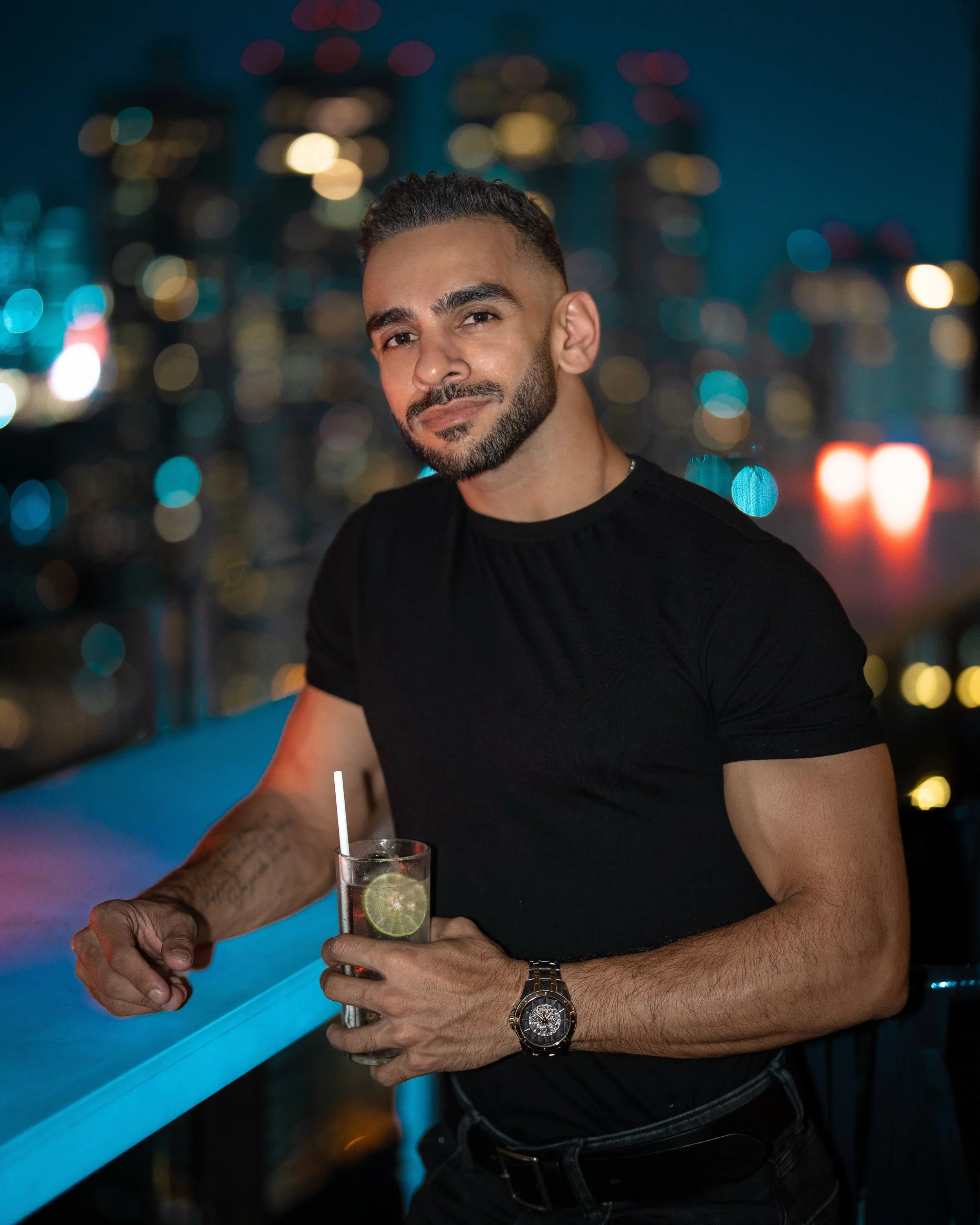 Kunal, a Bangkok photographer, with a beard and short dark hair wearing a black shirt and a silver watch, holding a cocktail glass with lime slice and straw, standing at a bar with a city skyline and colorful bokeh lights in the background at night.