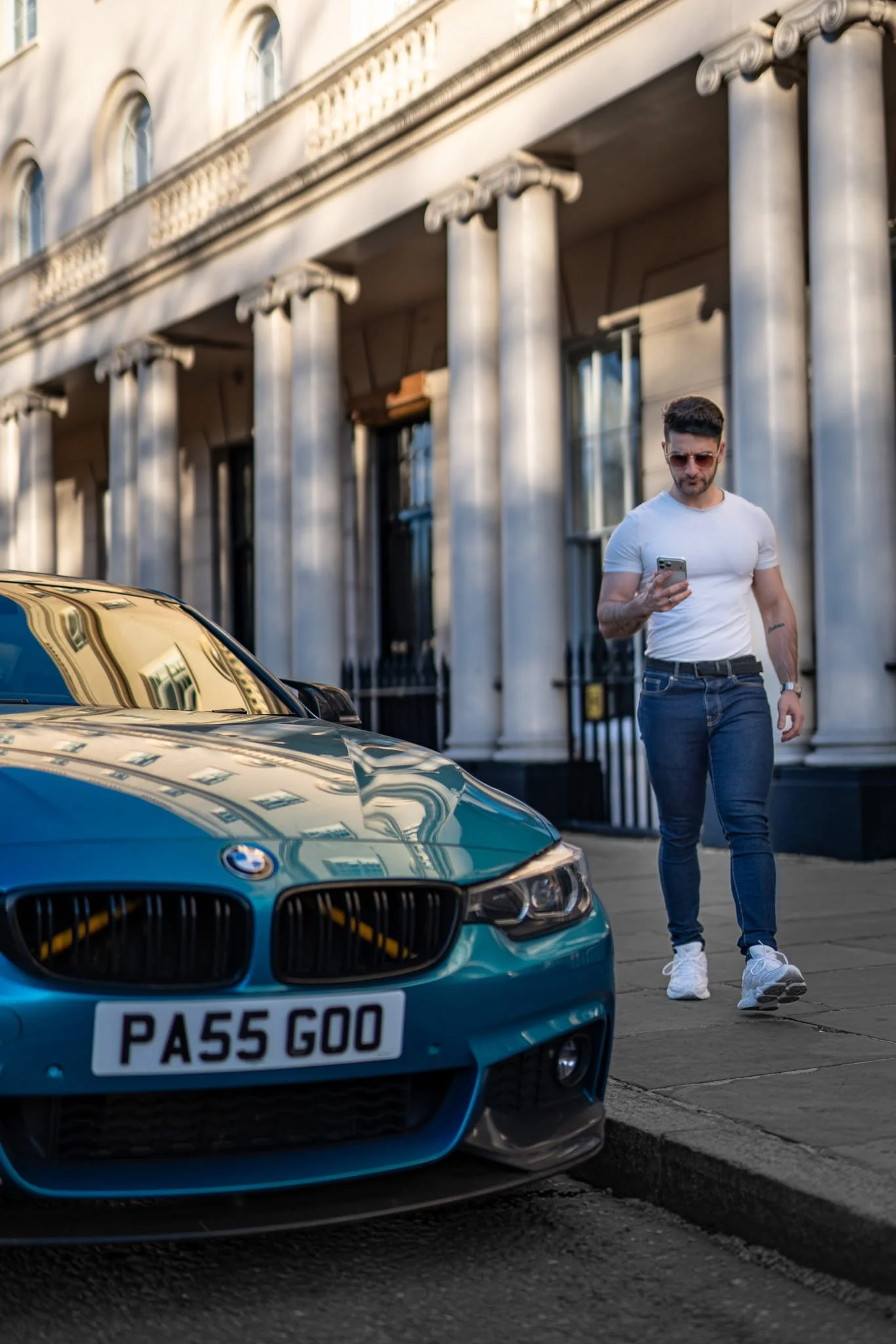 A man with sunglasses and a white t-shirt walking on a city sidewalk next to a blue BMW car with a license plate reading 'PA55 GOO'. The background shows a building with classical columns and windows.