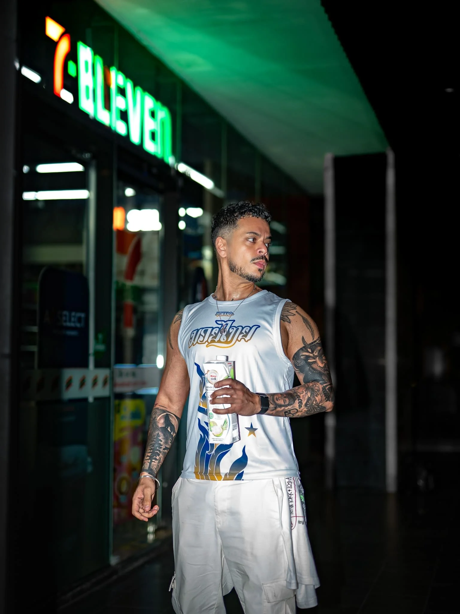 Young man with tattoos on his arms and face, wearing a sleeveless sports jersey and white shorts, holding a drink carton, standing outside a convenience store at night.