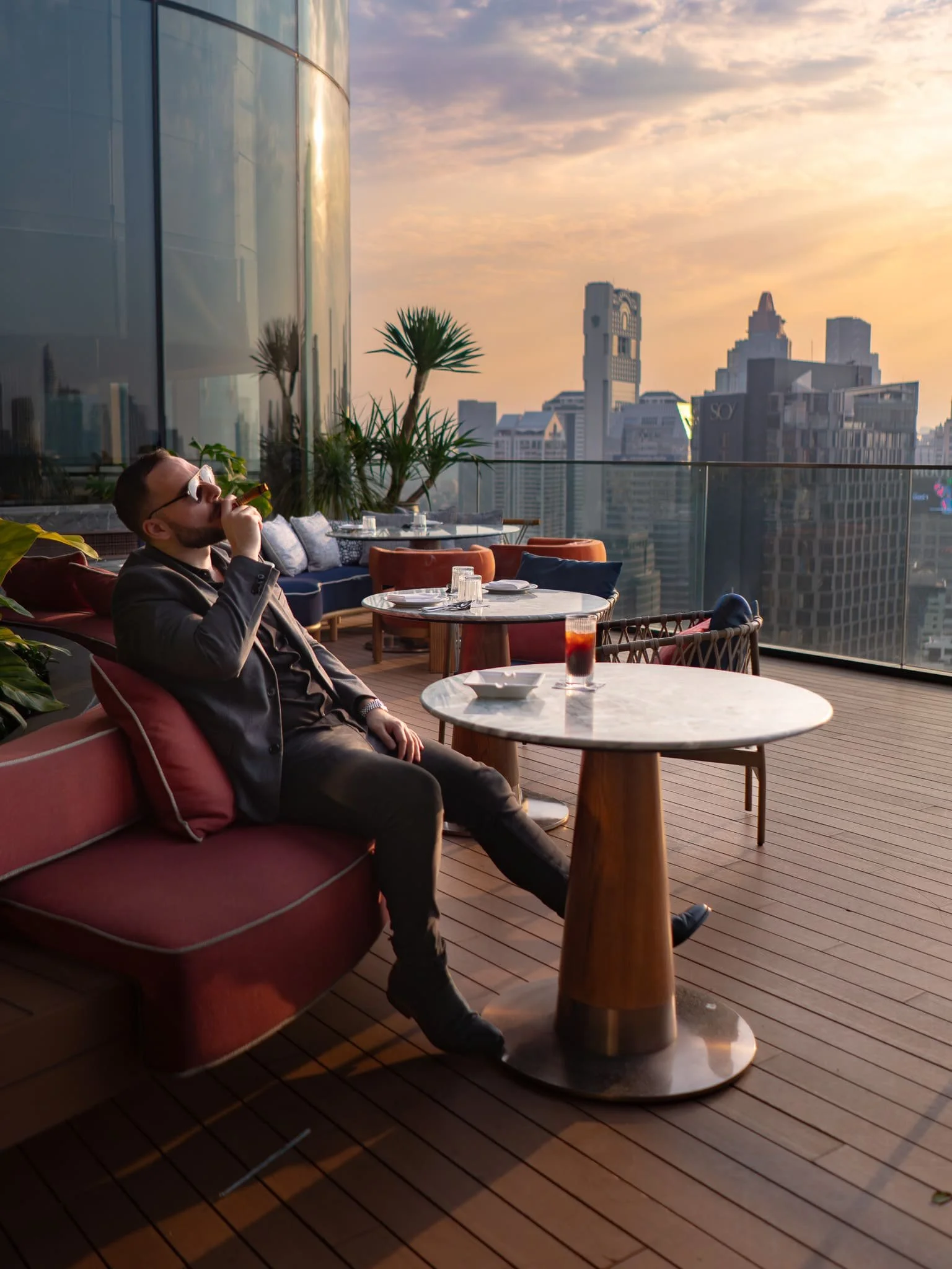 A man in sunglasses and a suit sitting on a red sofa on a rooftop terrace at sunset, drinking a beverage, with city skyscrapers and a cloudy sky in the background.