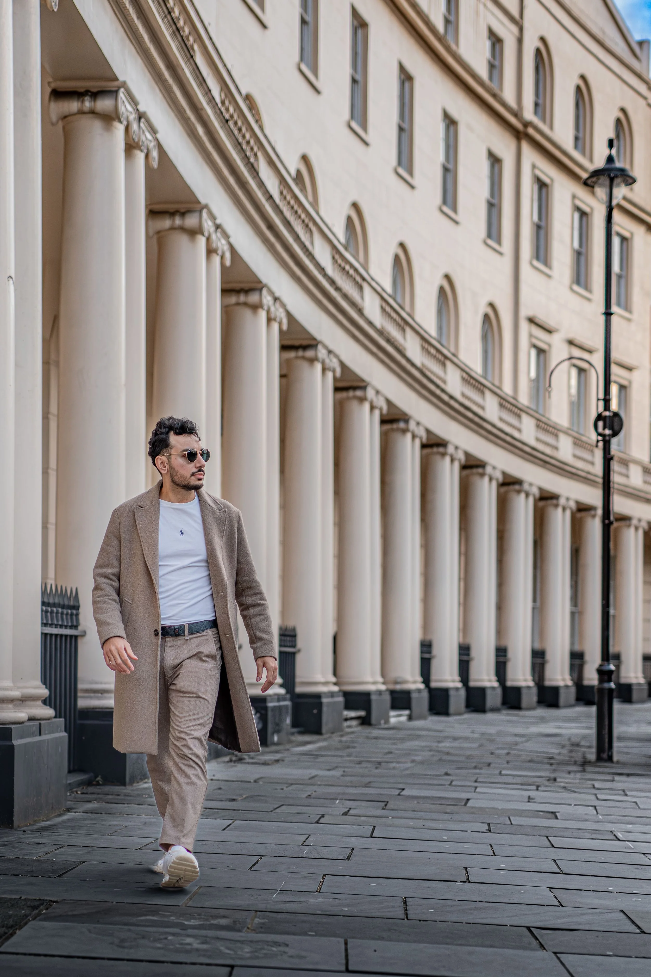 A man with dark hair and sunglasses walking along a city sidewalk with large columns and a cream-colored building in the background.
