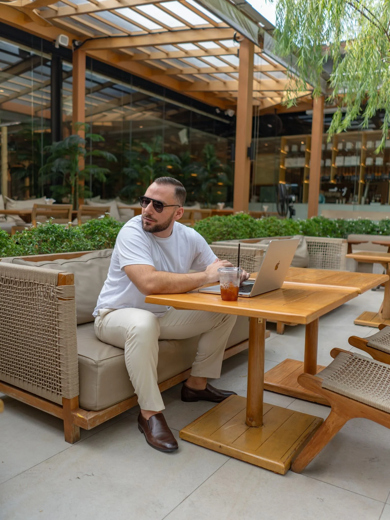 A man wearing sunglasses and a white t-shirt sitting at a wooden table with a laptop and a drink in a modern outdoor cafe with greenery and wooden decor.