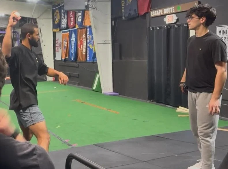 Two young men on a green indoor sports court, with flags hanging on the wall behind them. One man is wearing a black hoodie and shorts, gesturing with his hand, while the other man is standing facing him, wearing glasses, a black shirt, and gray sweatpants.