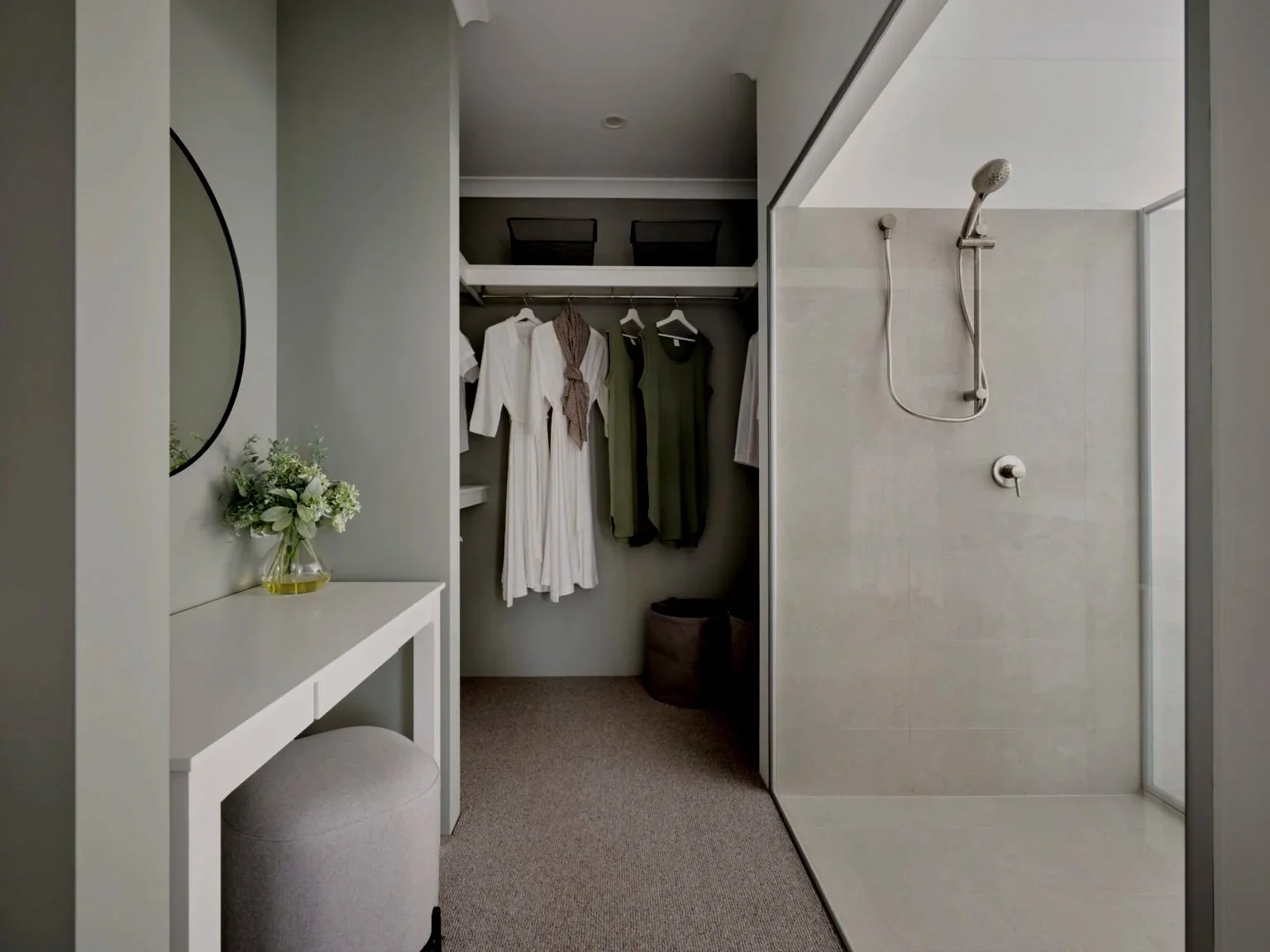 A walk-in closet with a small vanity table, a mirror, a vase of white flowers, and a cushioned stool. There is an open closet with hanging clothes and storage boxes. A glass-walled shower with a rainfall showerhead is visible to the right.