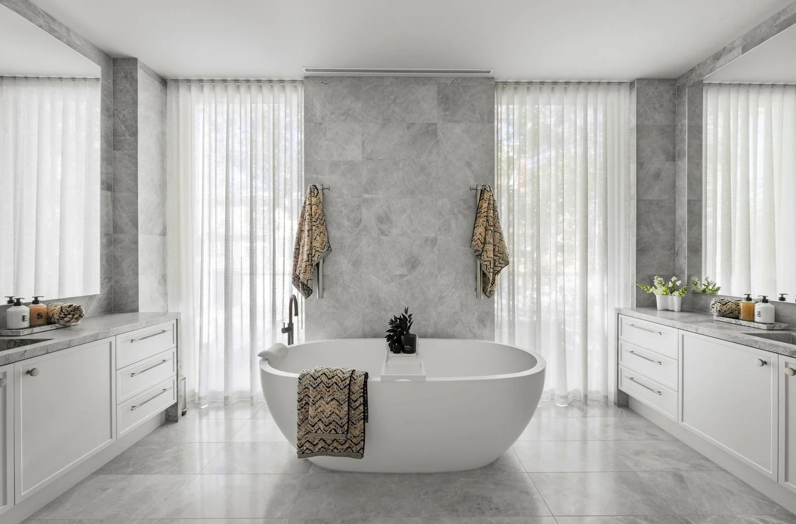 Modern bathroom with a freestanding oval bathtub in the center, flanked by two white vanities with marble countertops, large windows with sheer curtains, and decorative towels and plants.