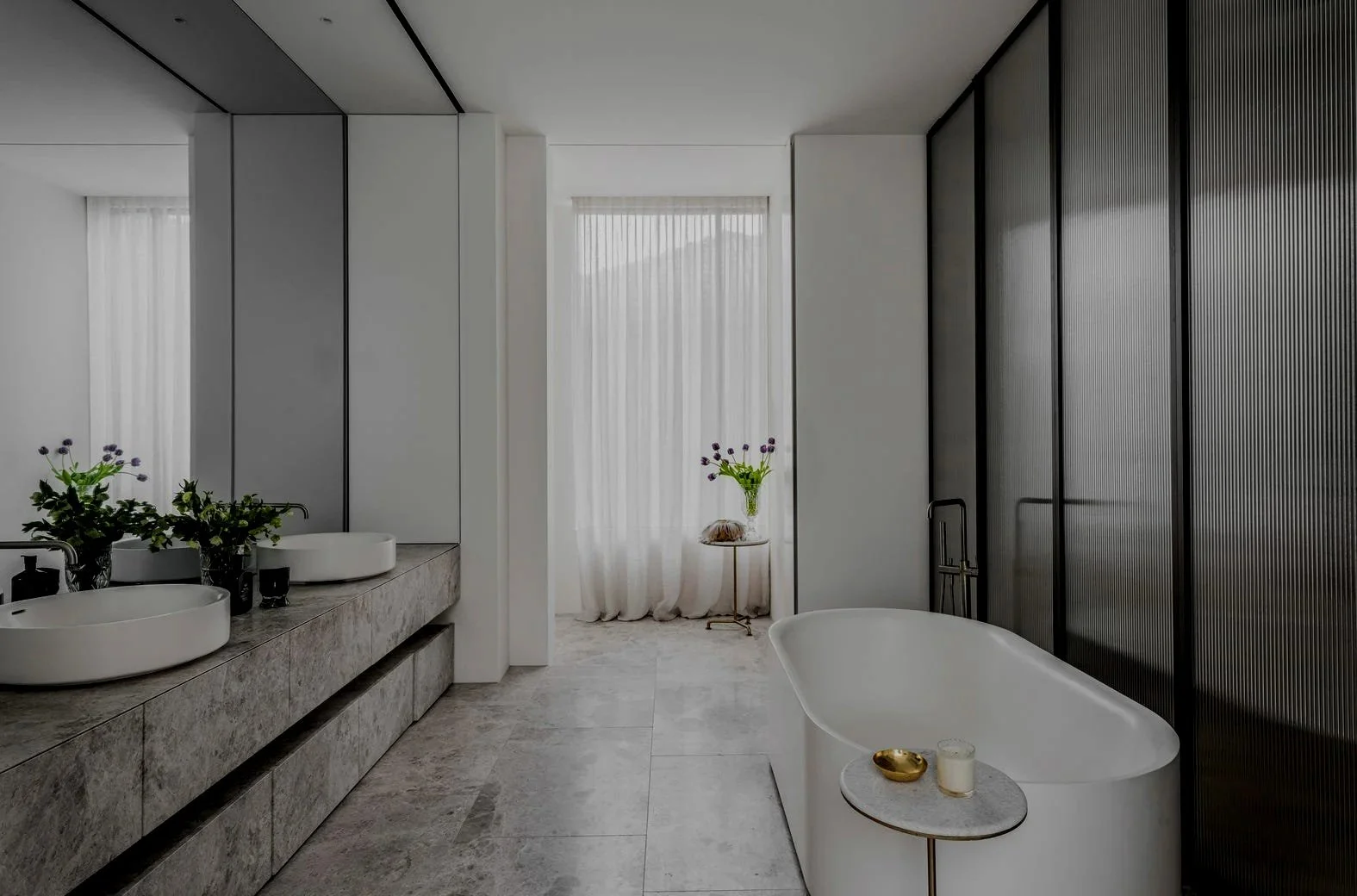 Modern bathroom with double sinks, marble countertop, white freestanding bathtub, and black lined glass cabinets, with sheer curtains and a small side table with a vase of flowers.