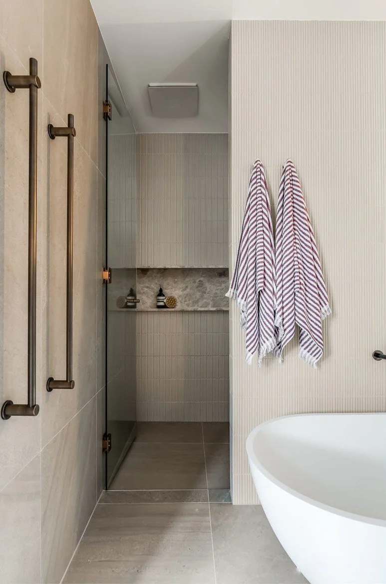 A modern bathroom with a corner shower that has a glass door, two striped towels hanging on the wall, a wall-mounted bathtub, and beige and textured wall and floor tiles.