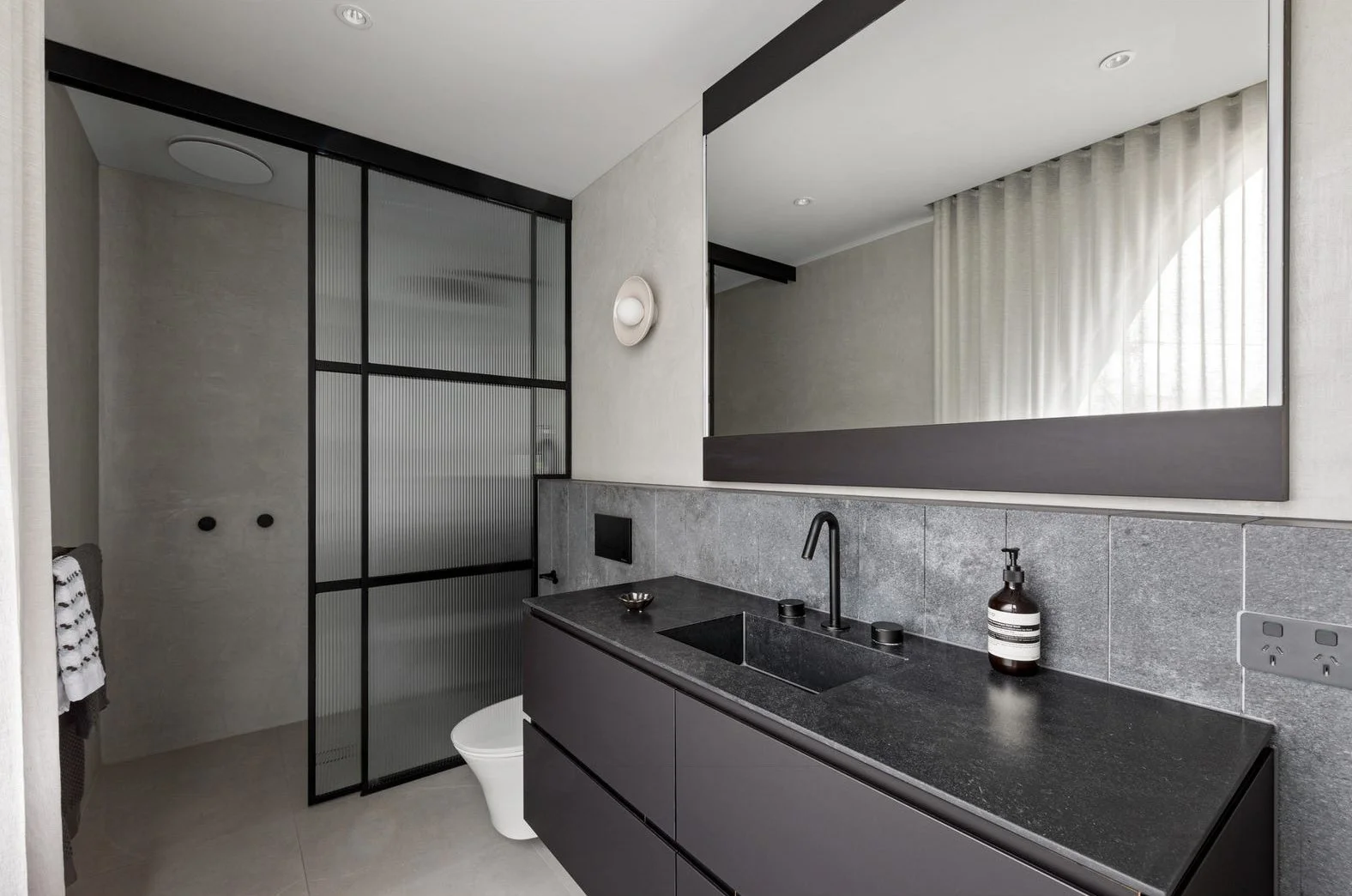 Modern bathroom with black vanity, dark countertop, large mirror, gray tiled wall, frosted glass partition, and minimal decor.