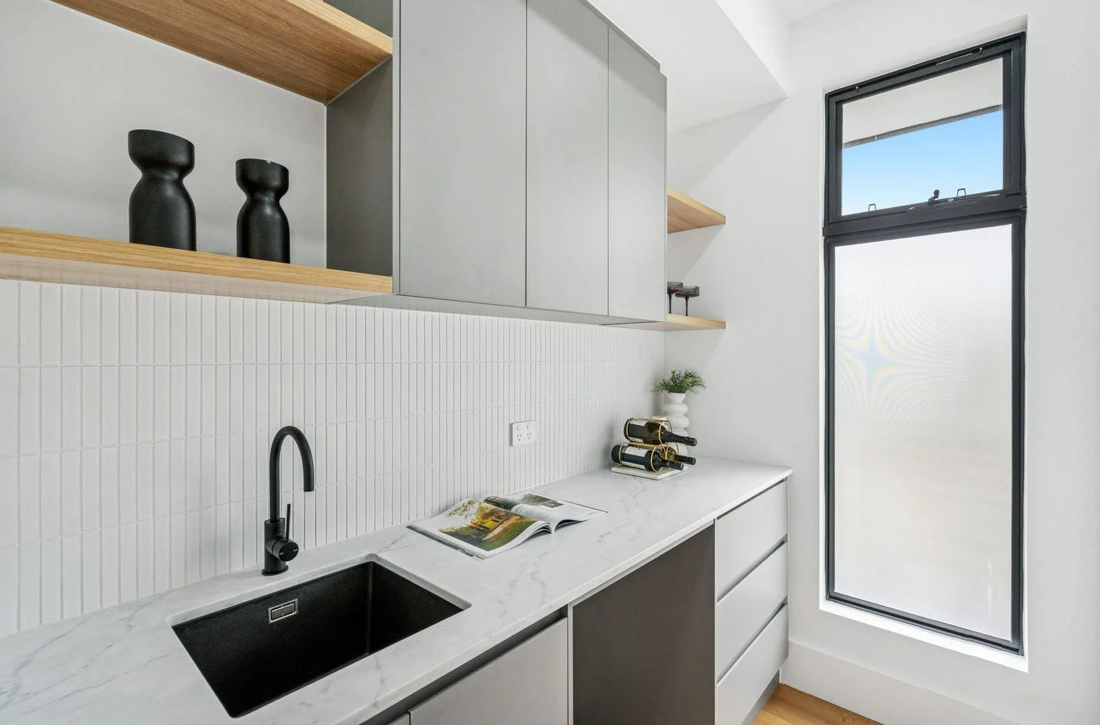 Modern kitchen with white marble countertop, black sink, black faucet, white tiled backsplash, gray cabinetry, wooden shelves, and a large window with black framing.