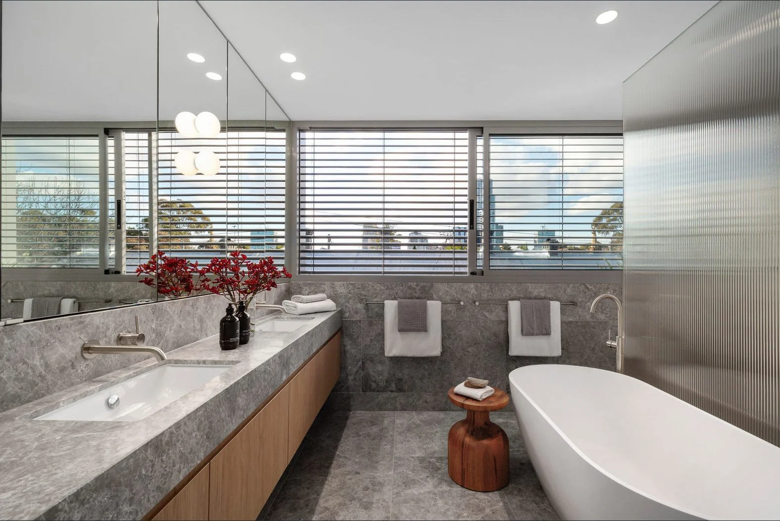 Modern bathroom with large windows, gray marble countertop with sinks, a freestanding bathtub, towels, and decorative red flowers.