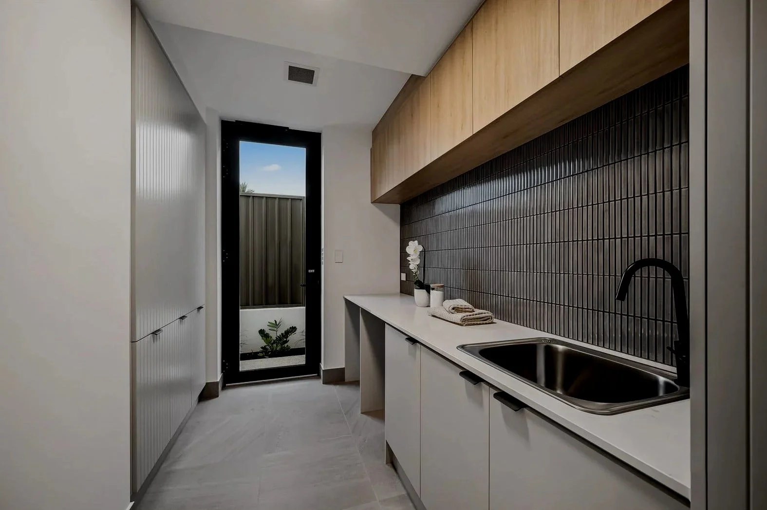 Modern kitchen with black tiled backsplash, white cabinetry, and a single sink. A black-framed glass door leads outside, with a small plant visible.