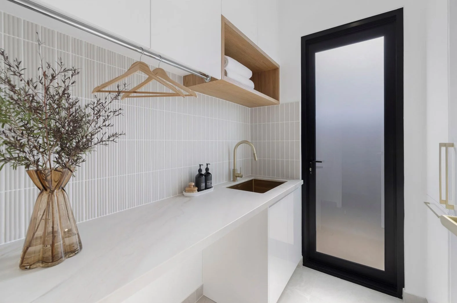 Modern laundry room with white countertops, a gold faucet, a wooden shelf with towels, a frosted glass door with black frame, and decorative items like a vase with eucalyptus and soap dispensers.