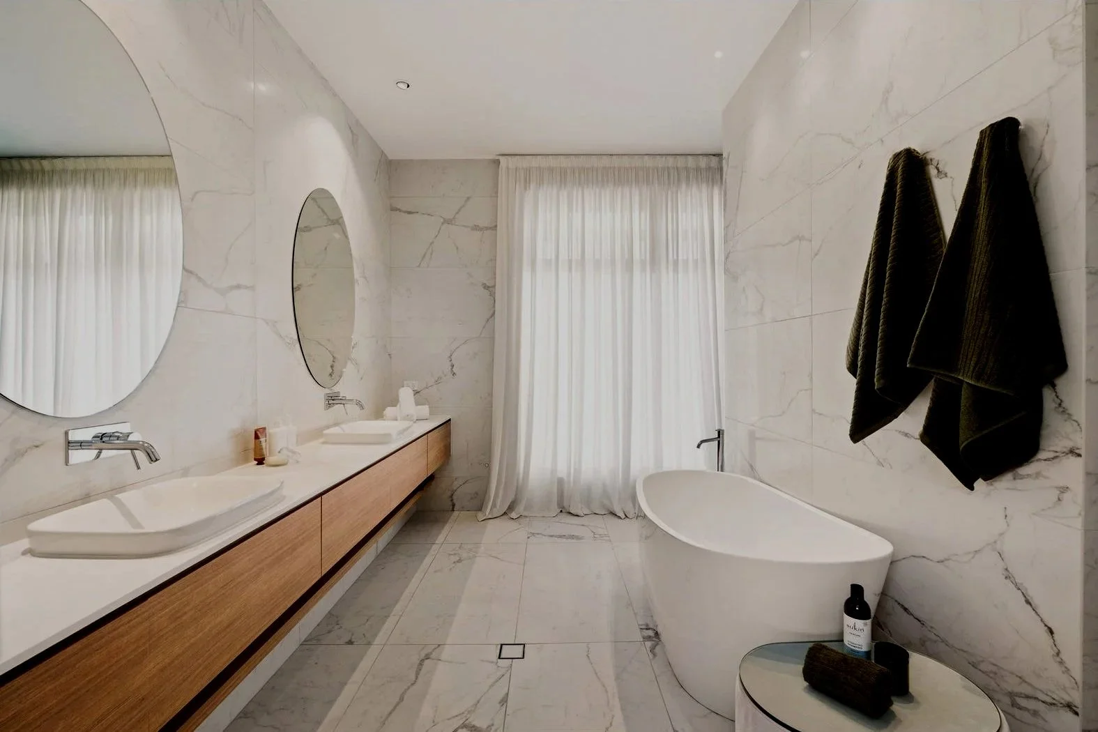Luxury bathroom with double vanity sinks, large round mirrors, a freestanding bathtub, and black towels on wall