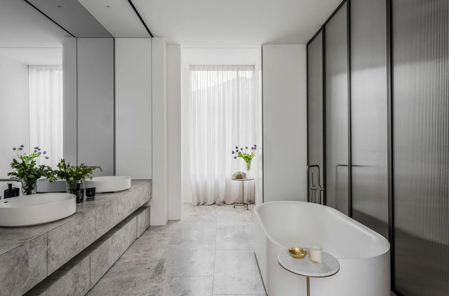 Modern bathroom with double sinks, large mirror, and hanging plants, featuring a freestanding bathtub and black and gray accents, with a window and white curtains.