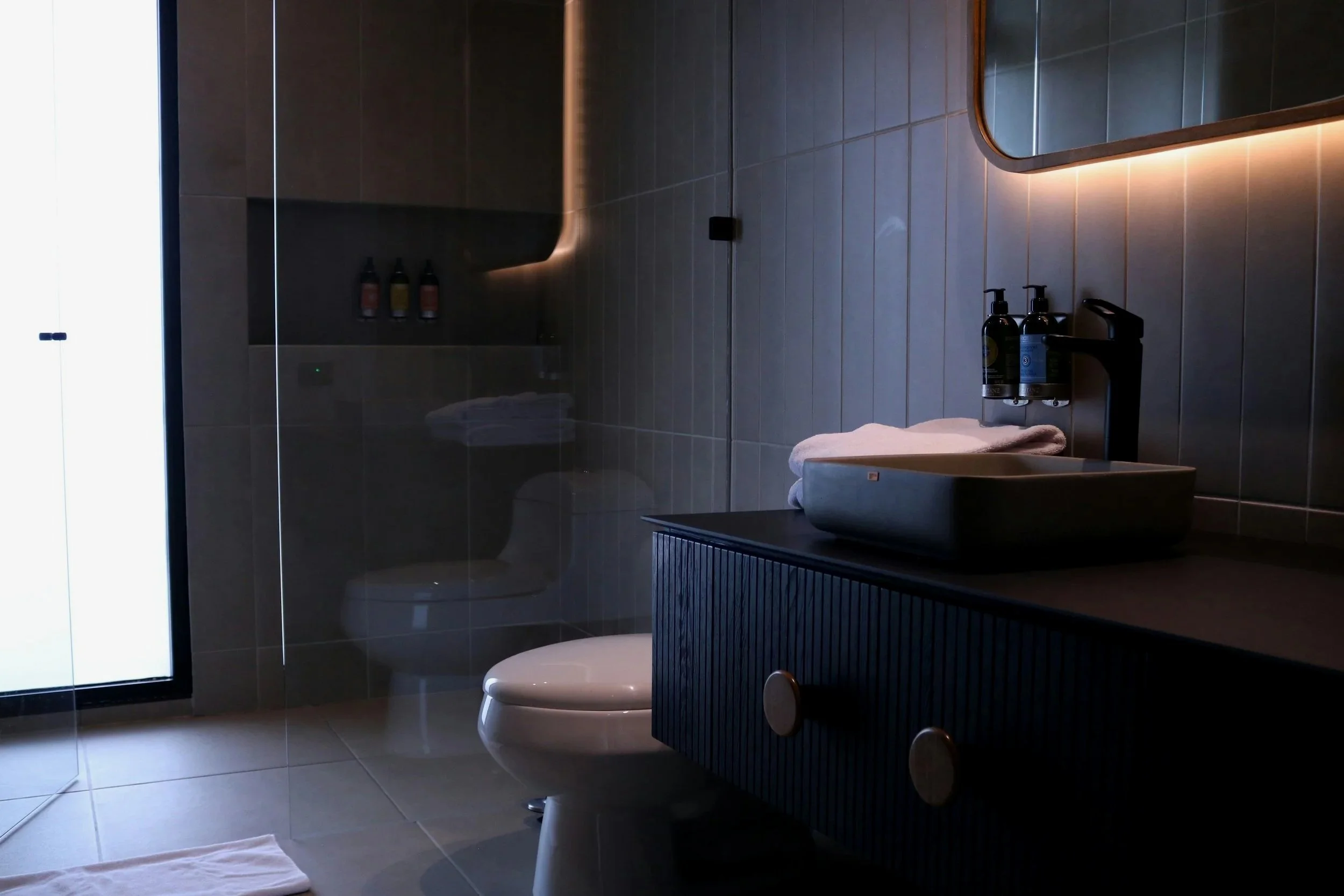 Modern bathroom with dark-colored vanity, a vessel sink, and a mirror with backlighting. Includes a toilet and toiletries on the wall. Soft lighting creates a cozy atmosphere.