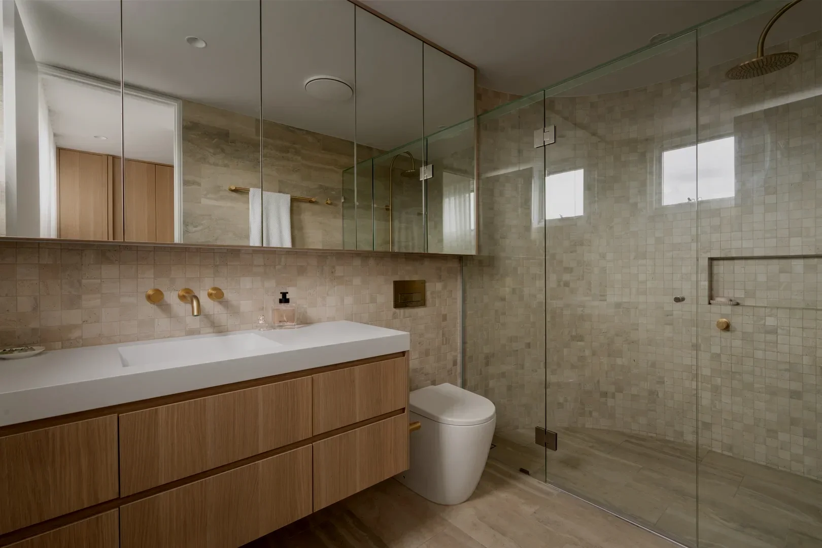 Modern bathroom with a shower area and a toilet. The shower has a black rain showerhead and a handheld shower, with a small built-in shelf. The bathroom has beige tiles and a black vent, with a toilet paper roll on the wall.
