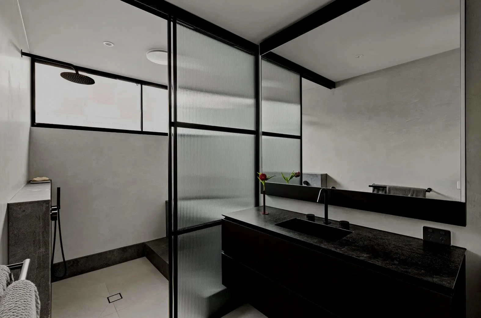 Modern bathroom with black and white color scheme, featuring a walk-in shower with a rain shower head, a black sink vanity with a large mirror, and a frosted glass partition.