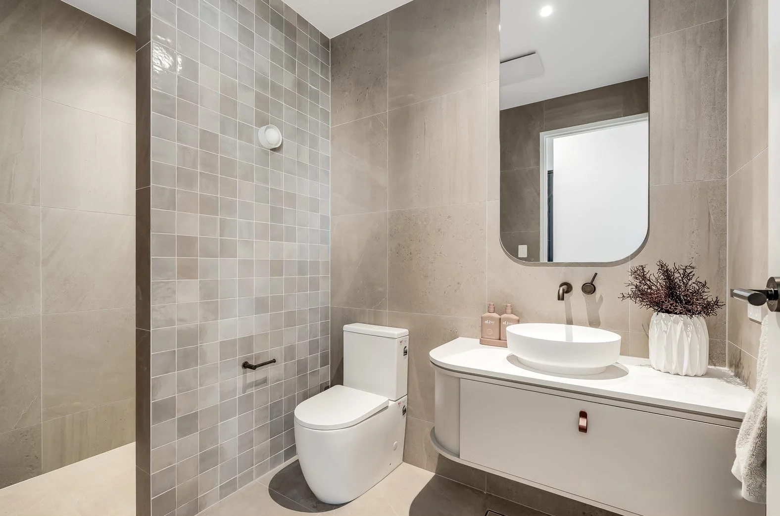 Modern bathroom with beige tiles, a white toilet, a curved vanity with a vessel sink, a round mirror, a vase with dried plants, and pink soap bottles.
