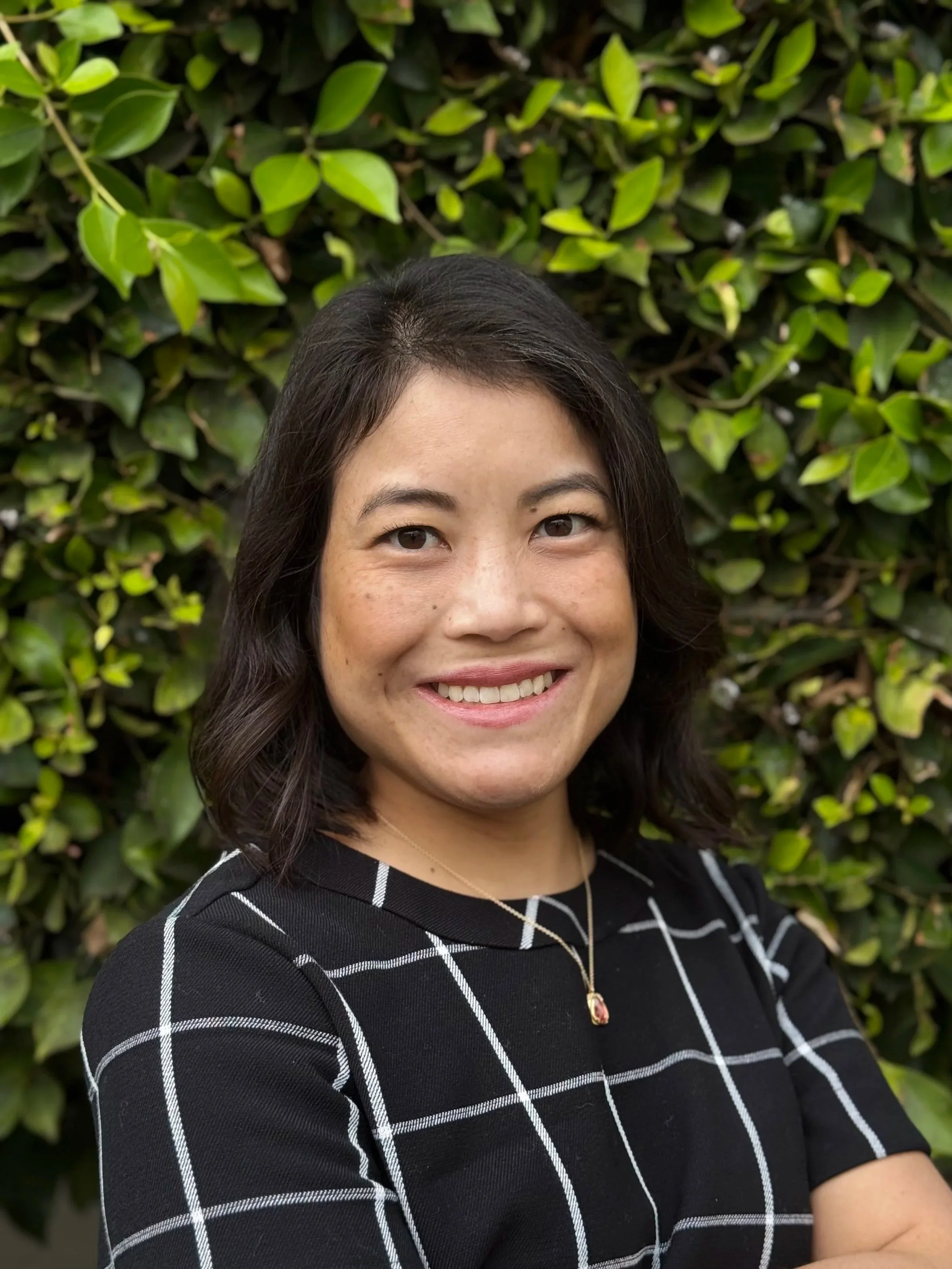 Dr. Minh-Chau Do smiling, standing in front of a green leafy hedge, wearing a black top with white checkered pattern and a gold necklace with a pendant.