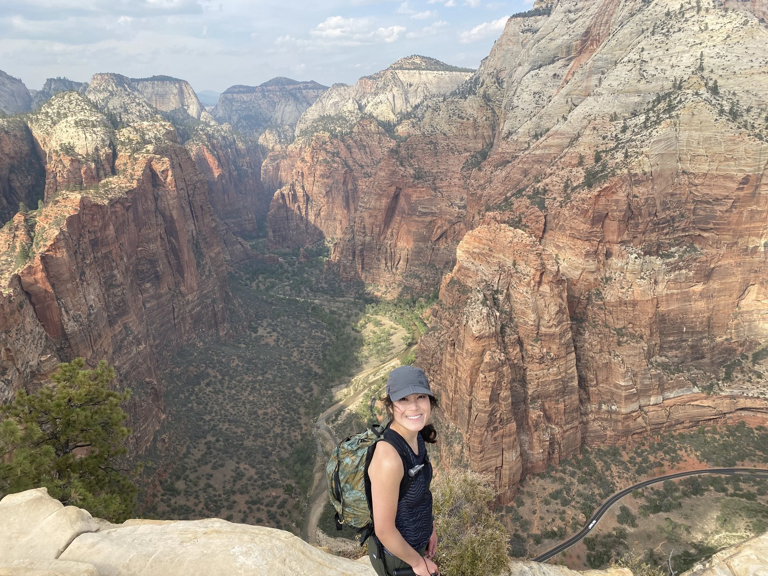 A smiling woman wearing a cap and backpack standing on a rocky ledge overlooking the canyon at Zion National Park in Utah during daytime.