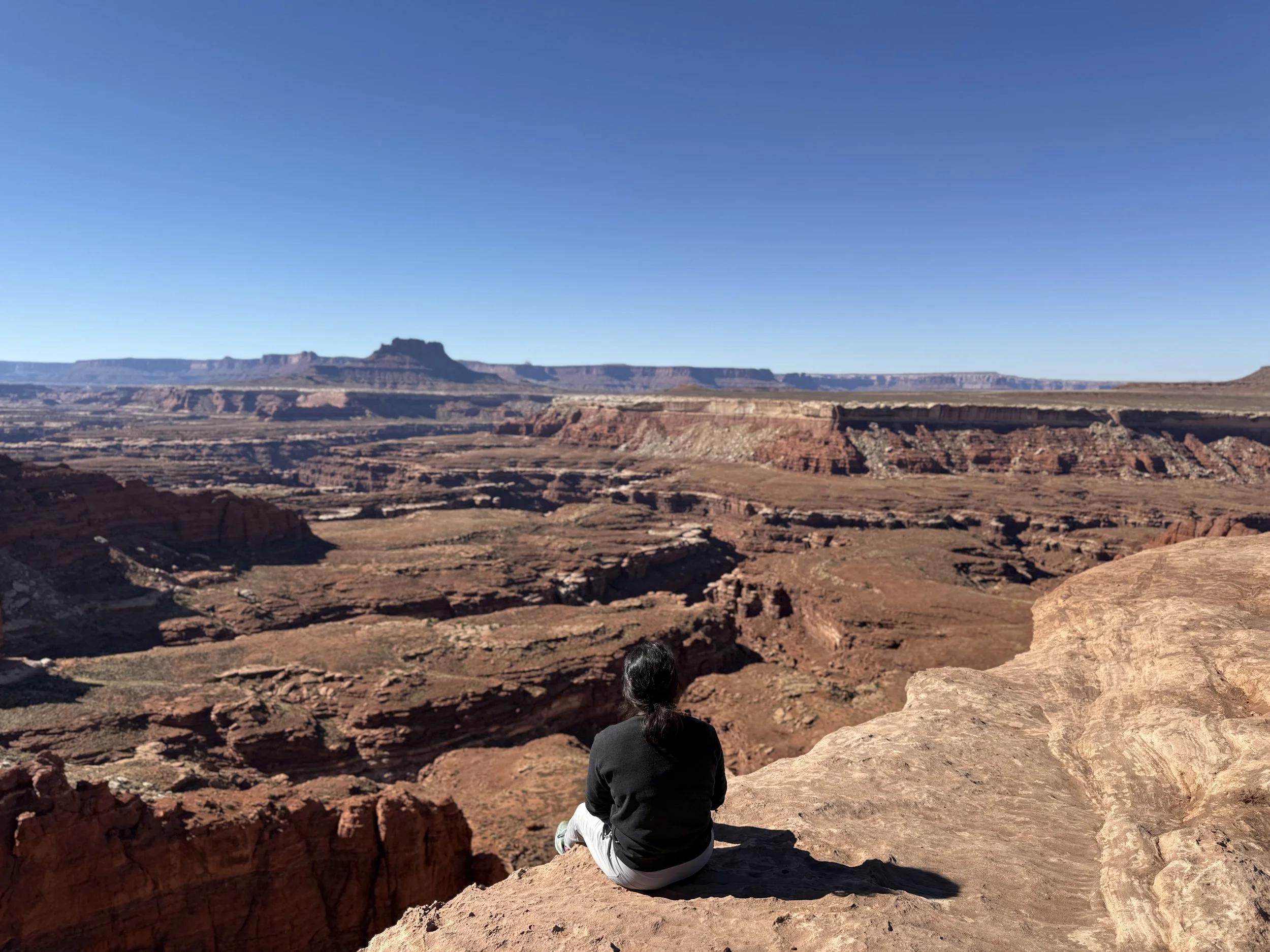 A person sitting on a rock ledge overlooking a vast red canyon with layered cliffs and mesas under a clear blue sky.