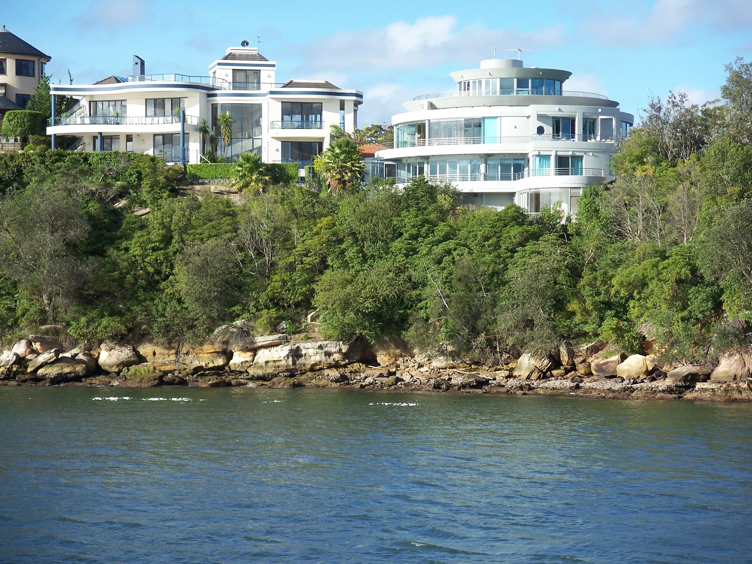 Modern white houses with large balconies situated on a lush green hillside by the water, connected by a private tunnel.