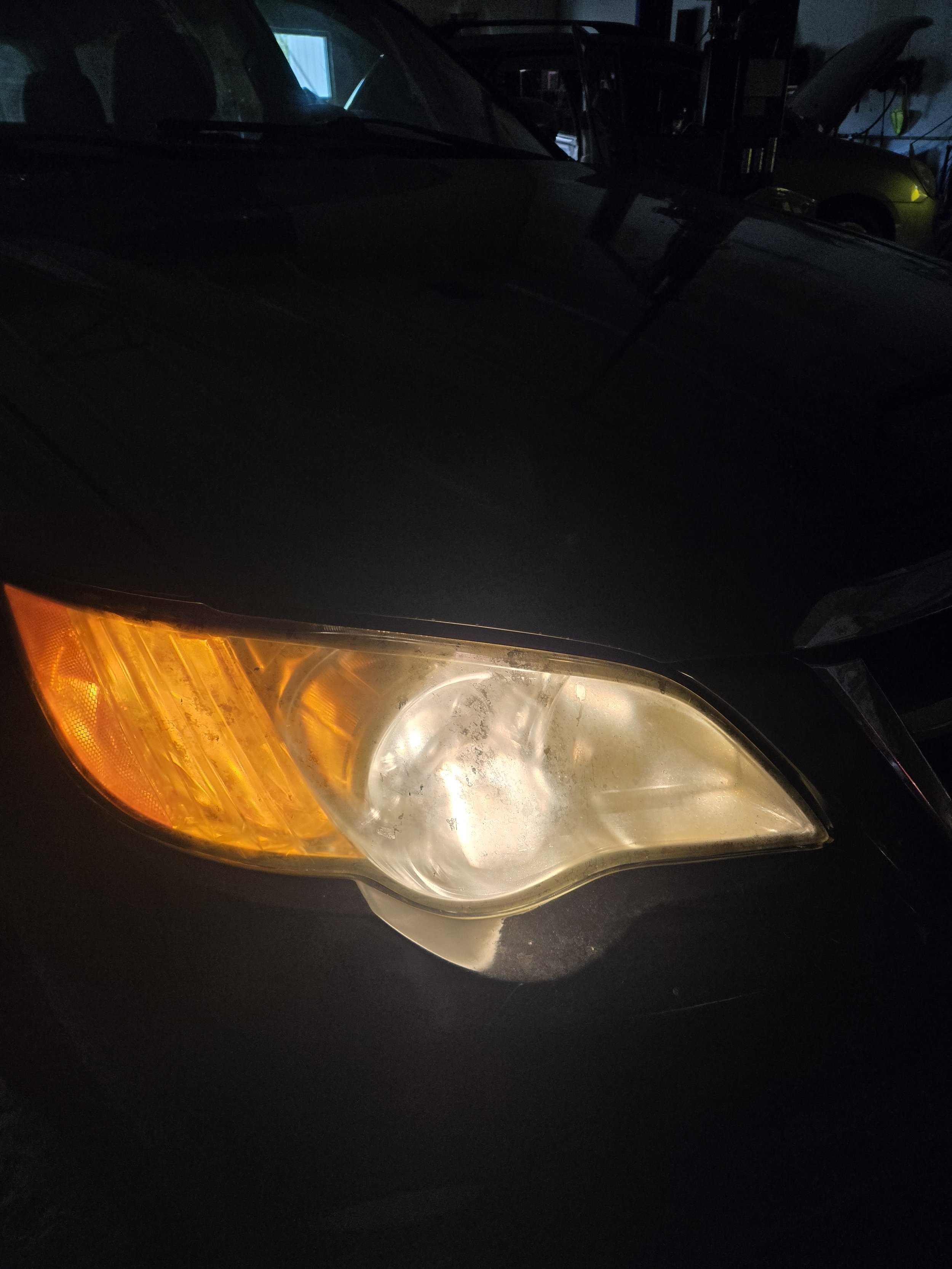 Close-up of a car headlight with an amber turn signal, showing signs of fogging and dirt, in a dimly lit garage.