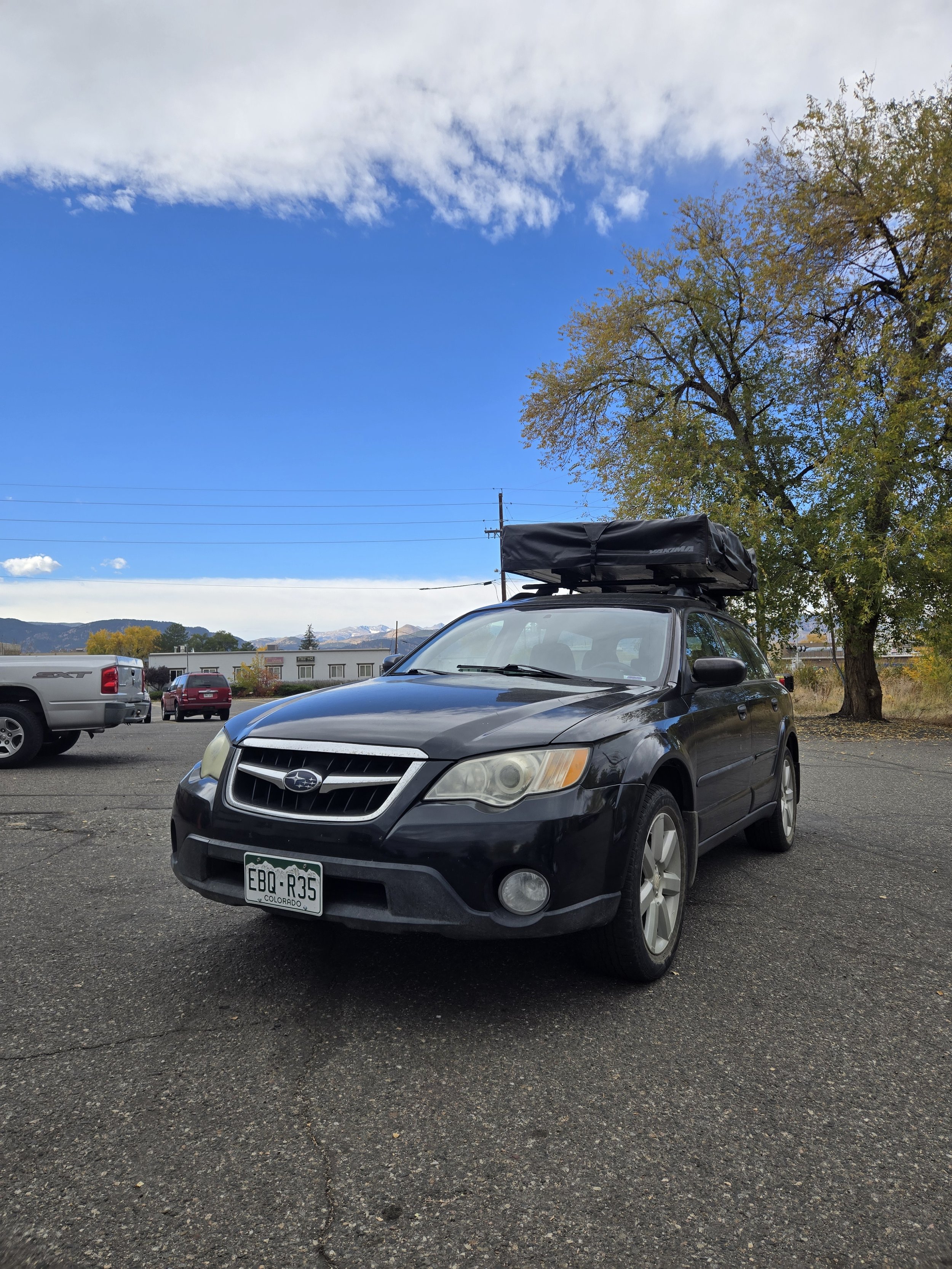 Black Subaru station wagon with gray tint windows, a roof cargo bag, parked on an asphalt lot under a partly cloudy sky with trees and mountains in the background.