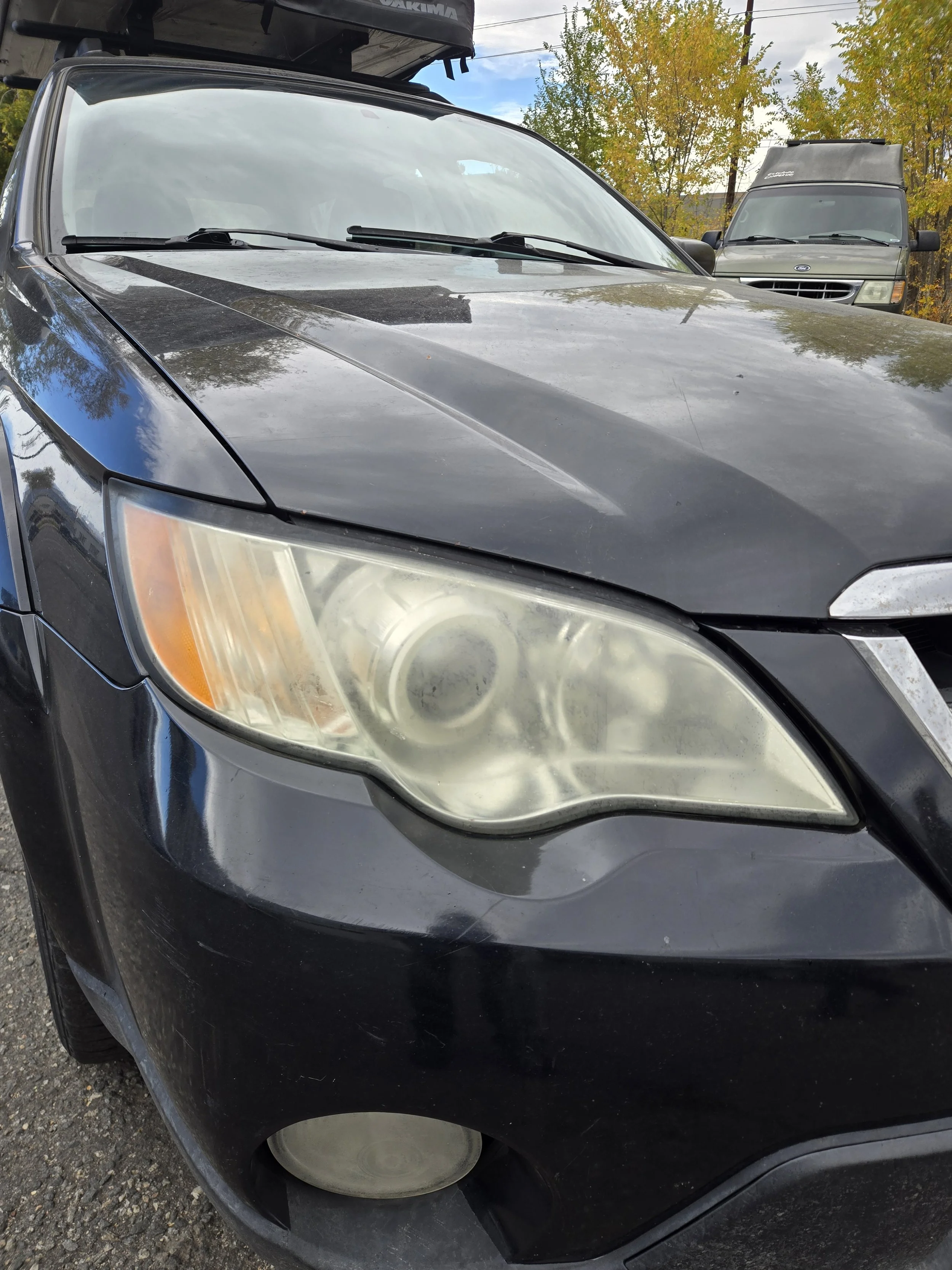 Close-up of a black car's front left headlight, showing foggy and cloudy condition, parked outdoors with another vehicle and trees in the background.