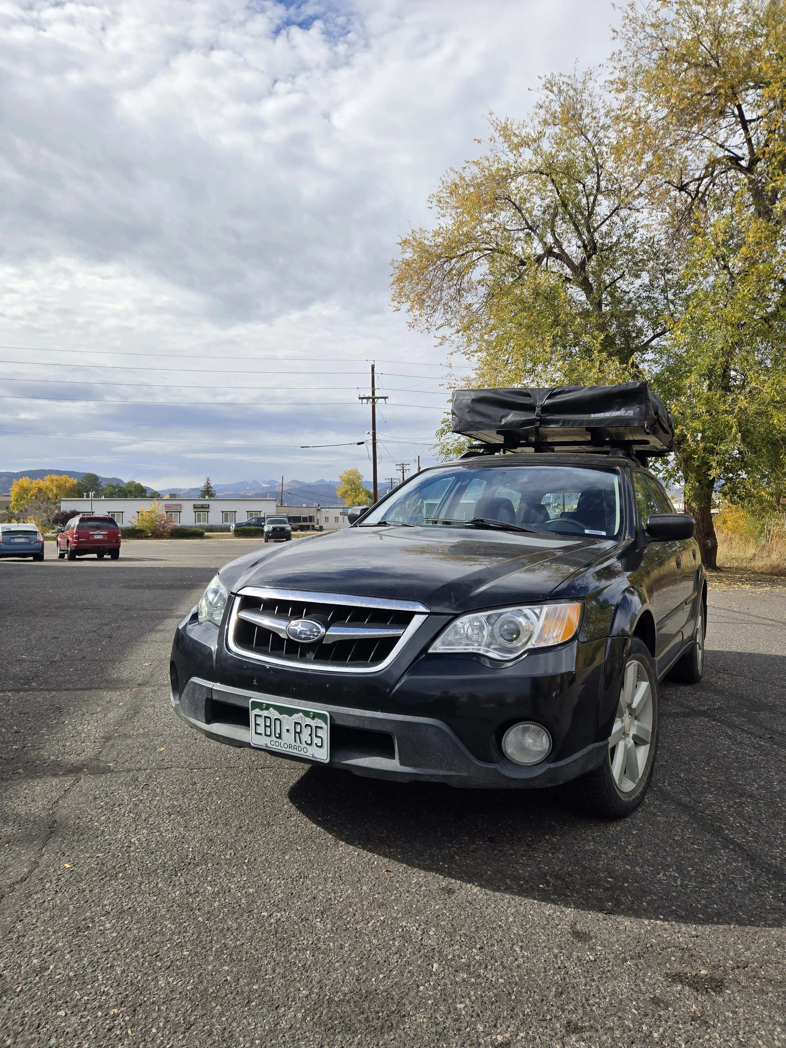Black Subaru car parked outdoors on a paved lot with a roof cargo box attached, trees with autumn foliage, and a cloudy sky in the background.