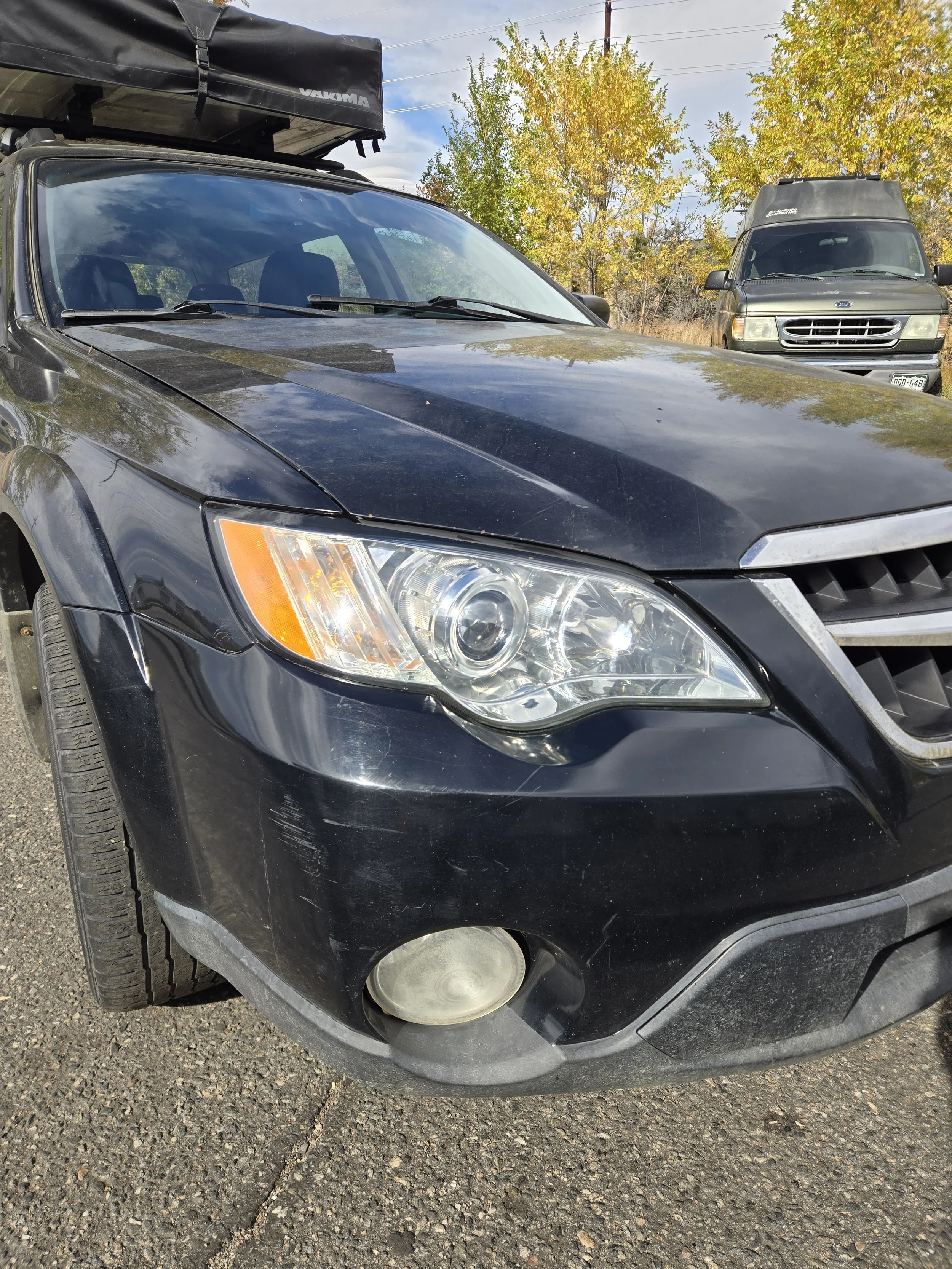 Black car with scratched front bumper and fog lights parked on asphalt, with yellow autumn trees and another vehicle in the background.