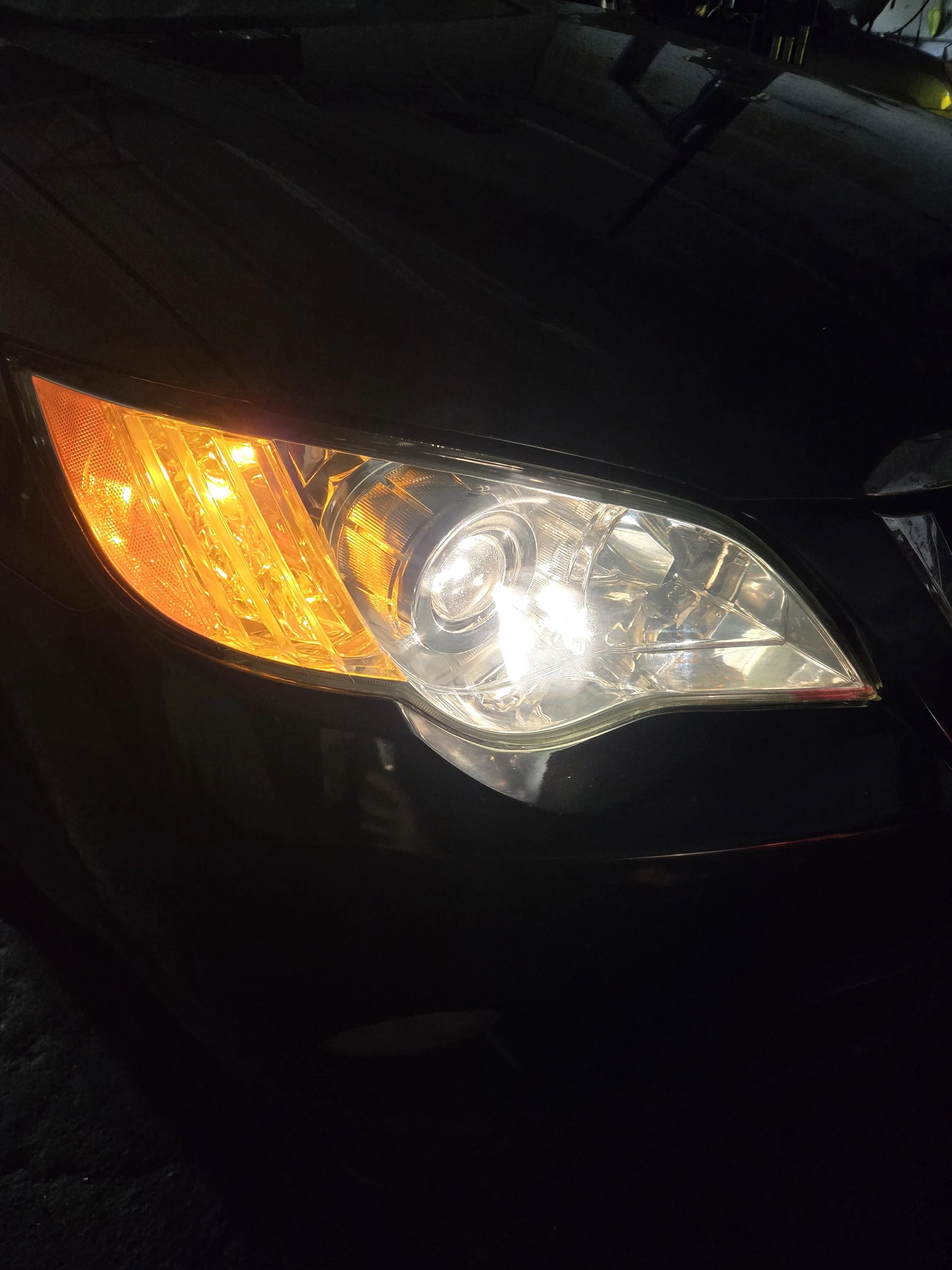 Close-up of a car's headlight and turn signal, with part of the black hood visible, taken in low light.