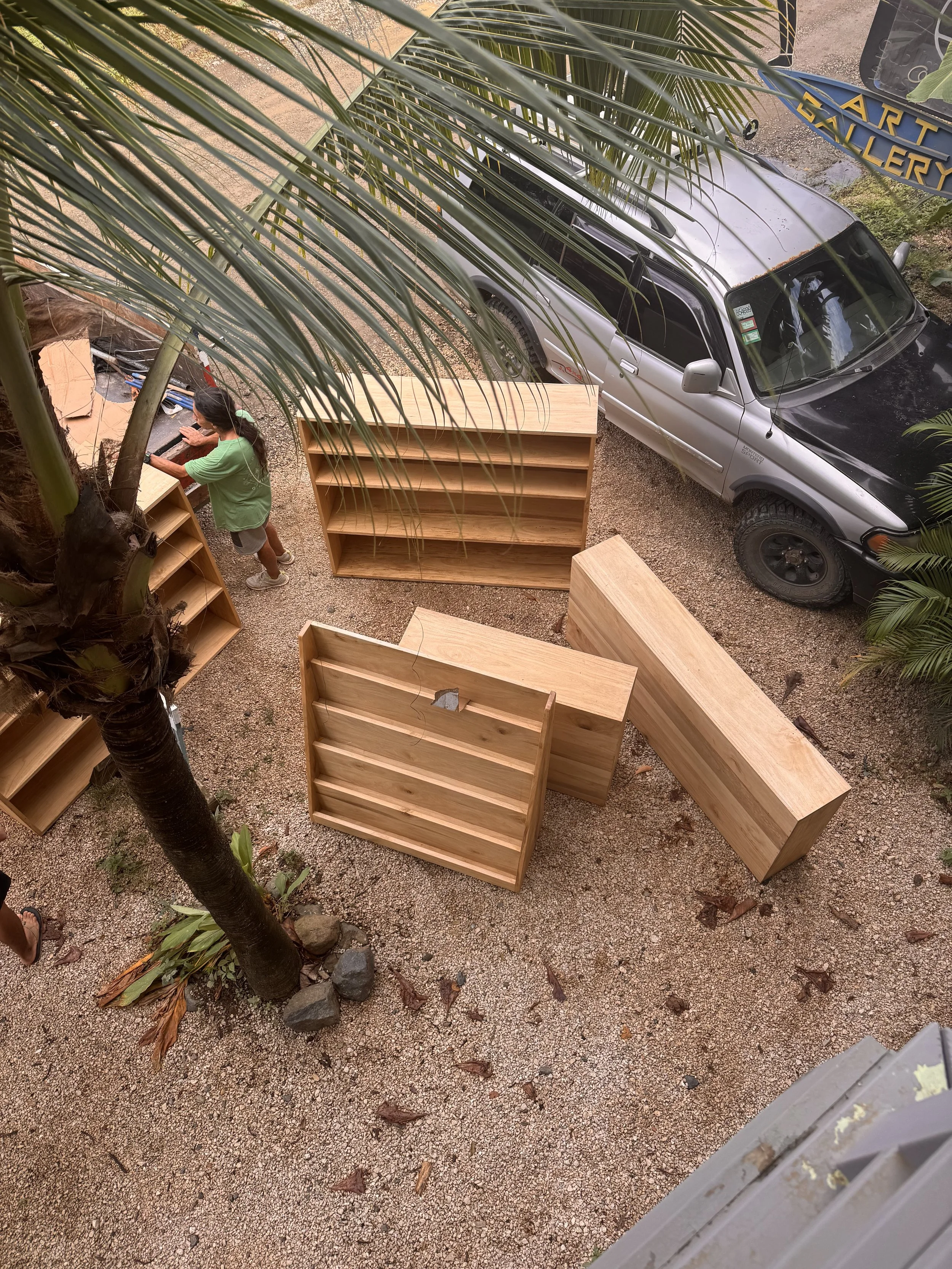 A woman building wooden shelves outside under a tree, with a silver SUV parked on a gravel driveway nearby.