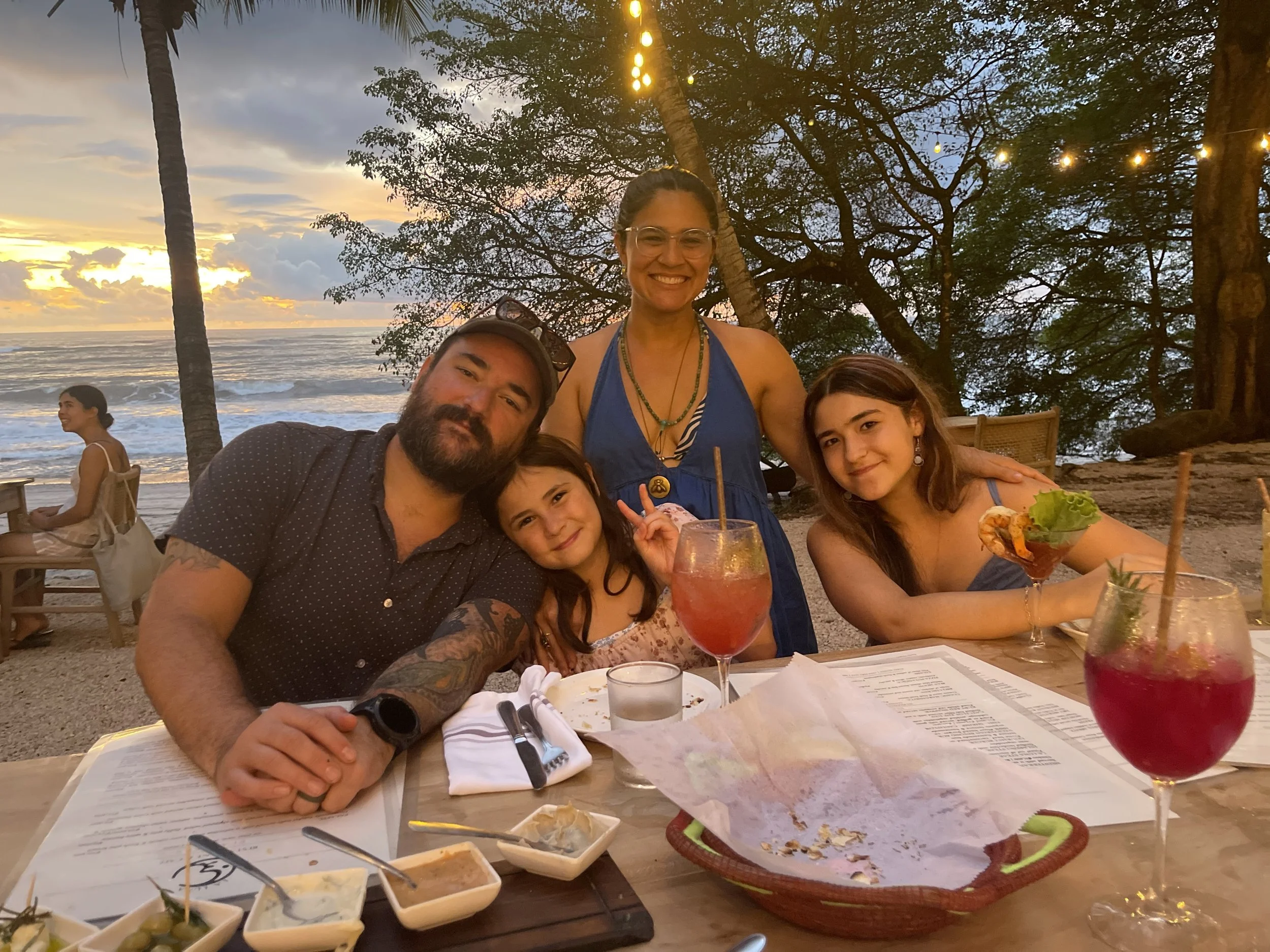 Group of four people enjoying dinner at a beachside restaurant during sunset, with drinks on the table and ocean view in the background.