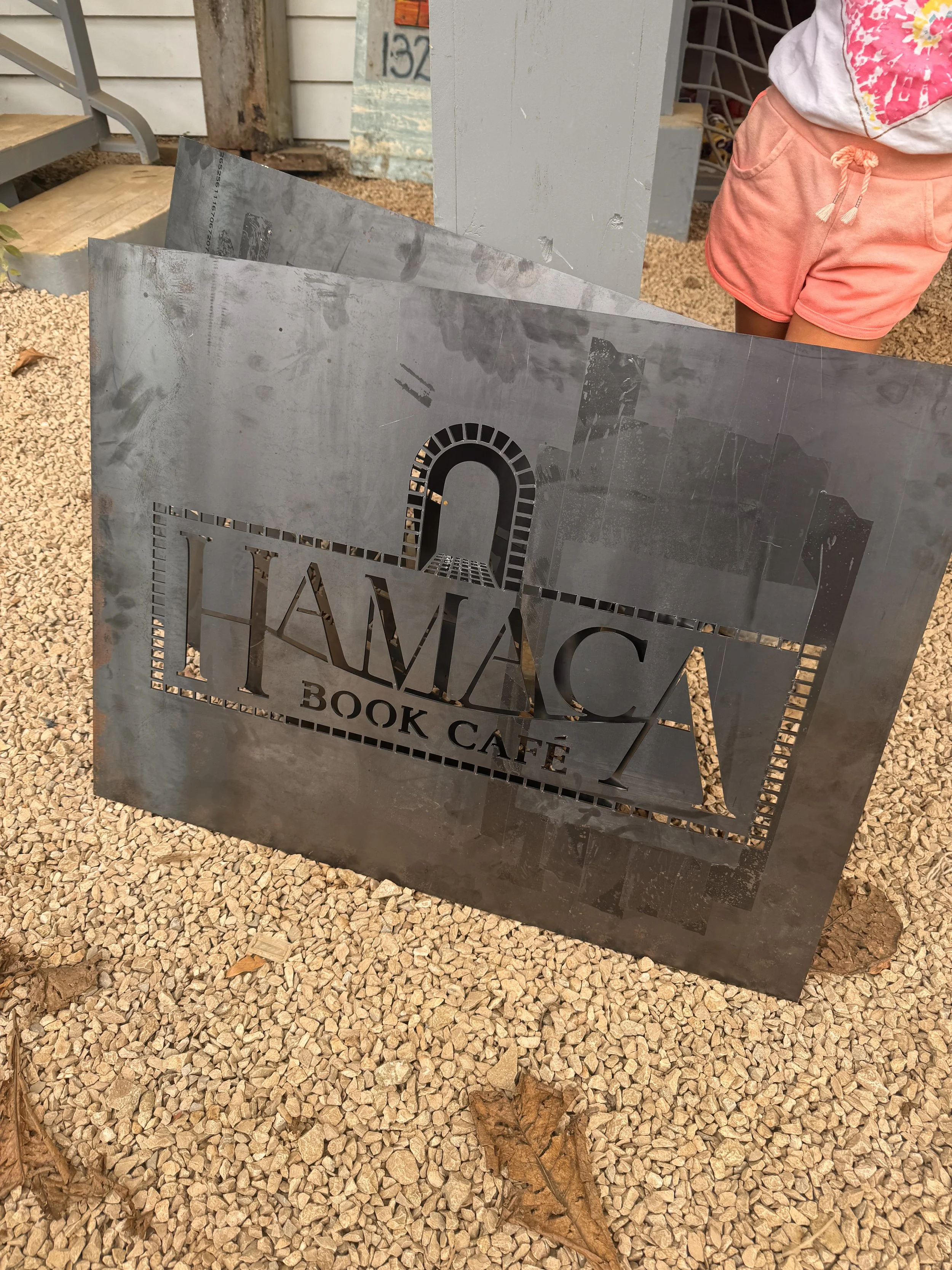 A metallic sign for 'HMAC Book Cafe' with cut-out text and a decorative archway design, standing on gravel ground with some fallen leaves, with a person wearing a pink outfit partially visible in the background.