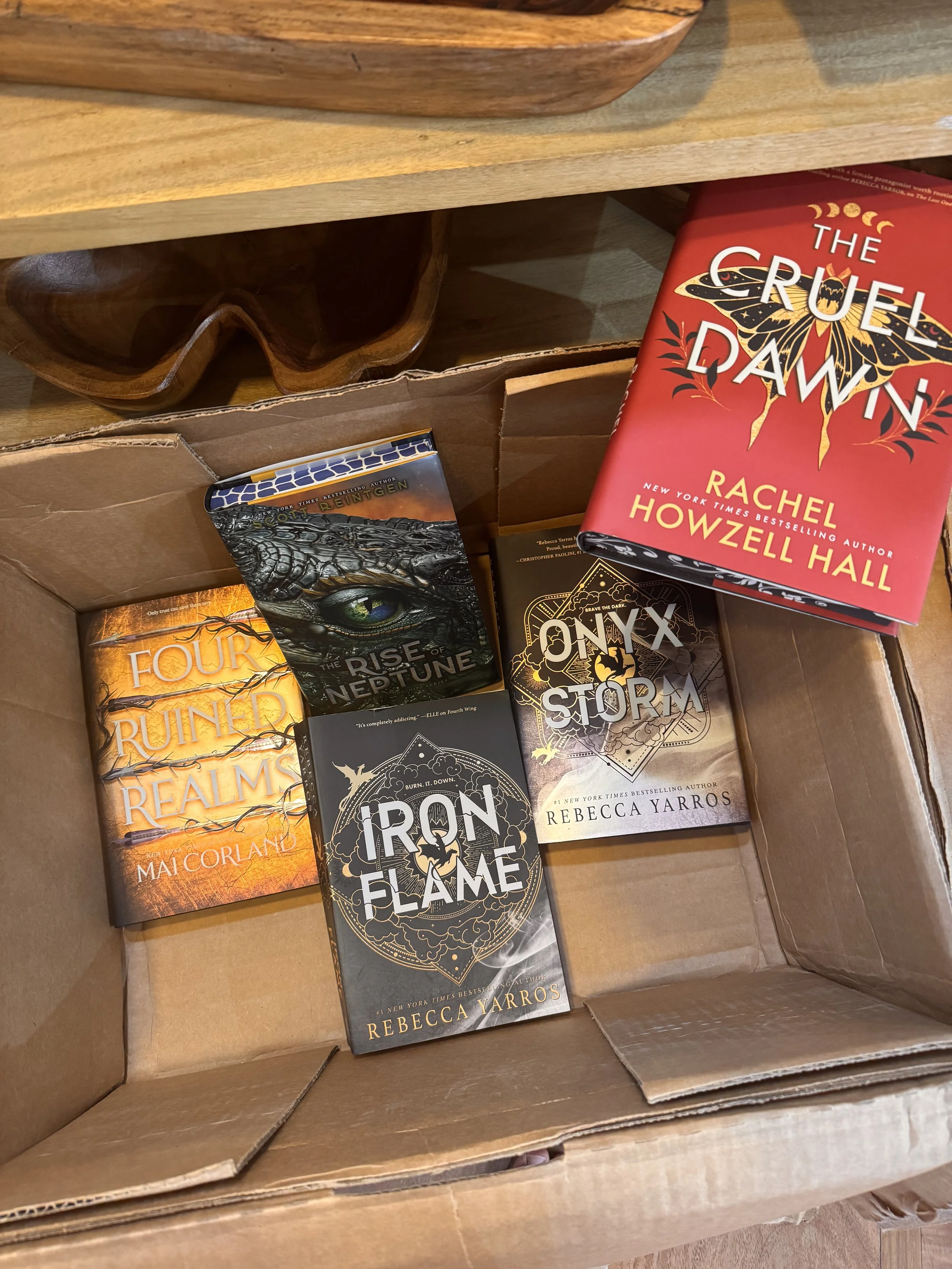 A cardboard box containing multiple fantasy and adventure novels, including 'Four Ruined Realms' by Mai Corland, 'The Rise of Neptune' by Rebecca Yarros, 'Iron Flame' by Rebecca Yarros, and 'Onyx Storm' by Rebecca Yarros.