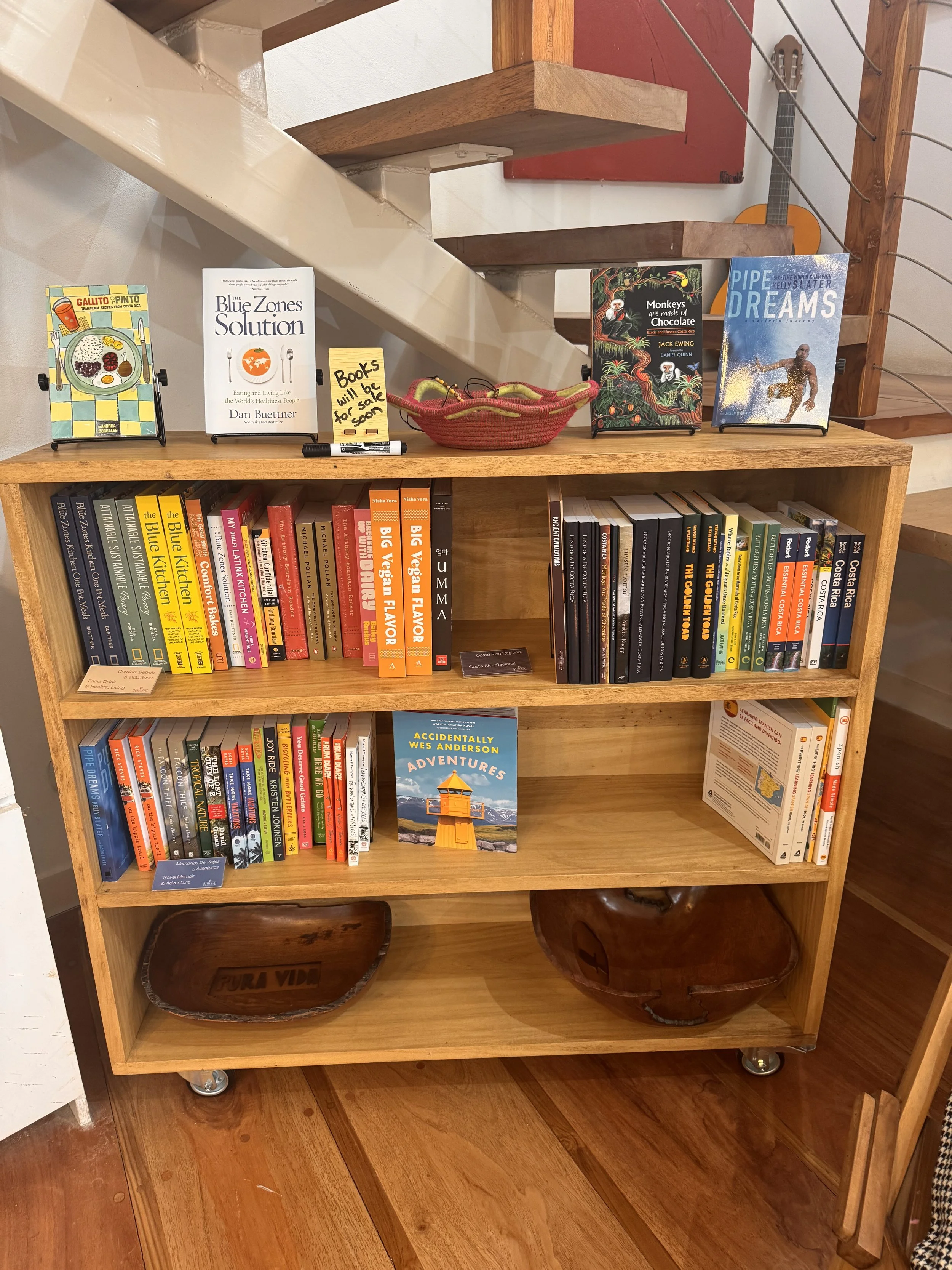 A wooden bookshelf on wheels displaying books, decorative bowls, and a sign that reads 'Books will be for sale' in a room with wooden floors and stairs in the background.