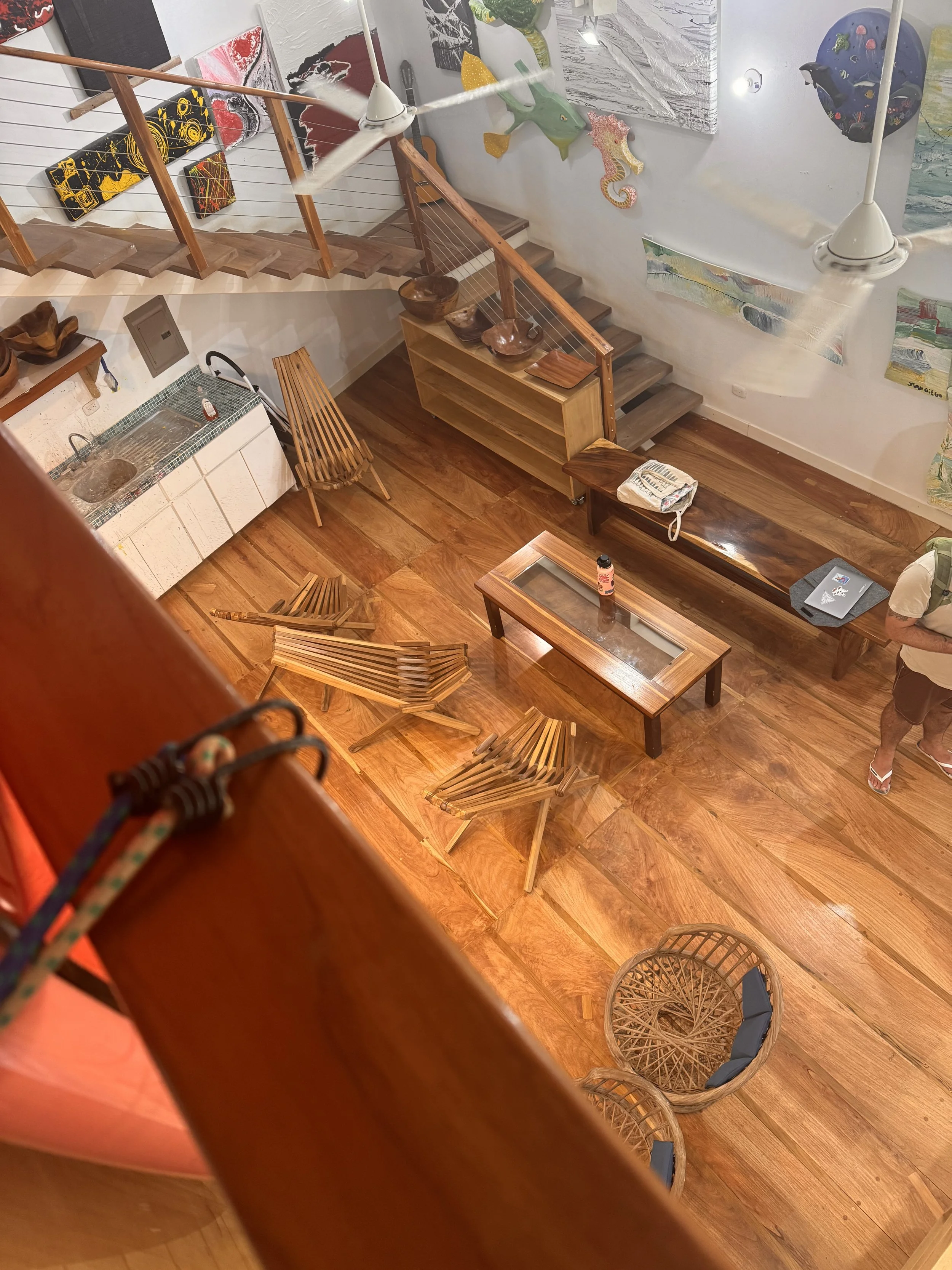 Interior view of a room with wooden flooring, wooden chairs, a bench, a glass-topped coffee table, and wall art including paintings of sea creatures and abstract designs. A person with a laptop and a water bottle on the table is visible.