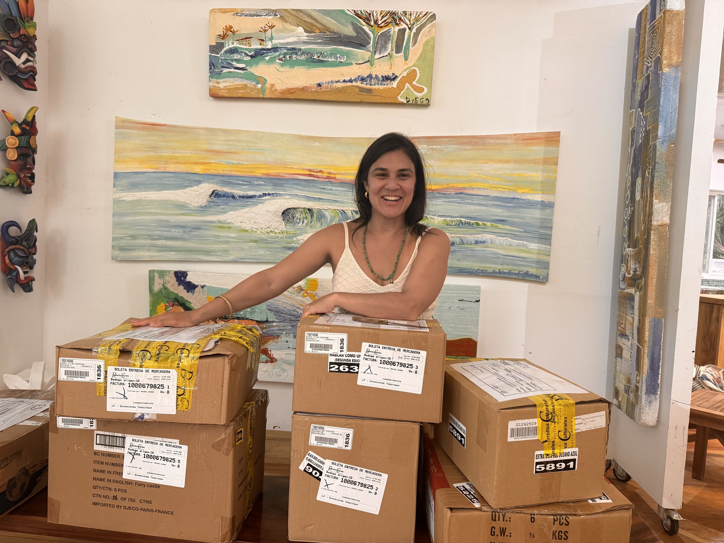 A woman smiling and standing behind a table with several cardboard boxes, in an art gallery or studio with colorful paintings of ocean waves and abstract art on the wall behind her.