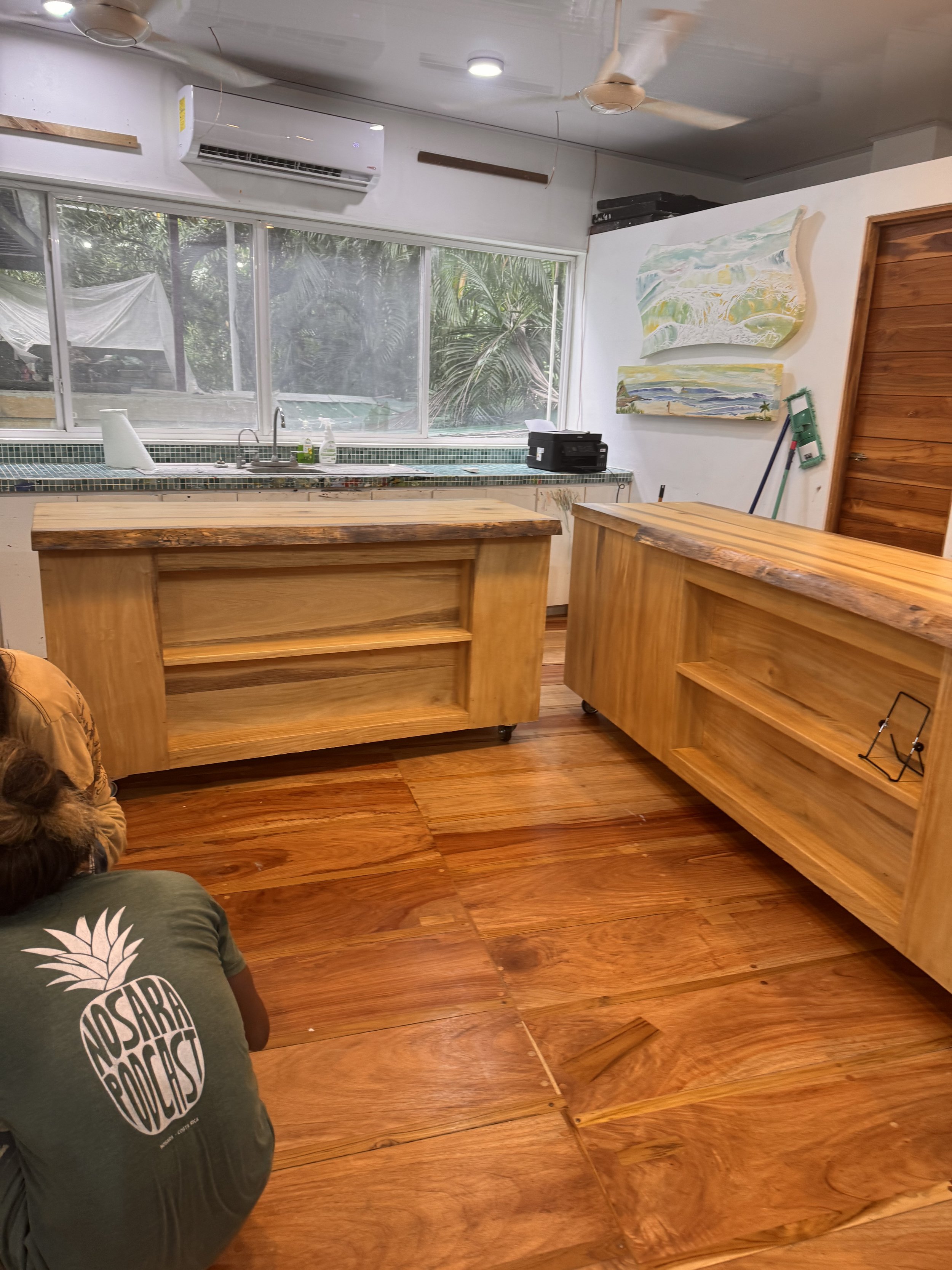Interior of a room with wooden flooring and wooden furniture, a kitchen sink in front of large windows, with paintings on the wall, a person wearing a green t-shirt with 'Nosara Podcast' on the back is kneeling on the floor.