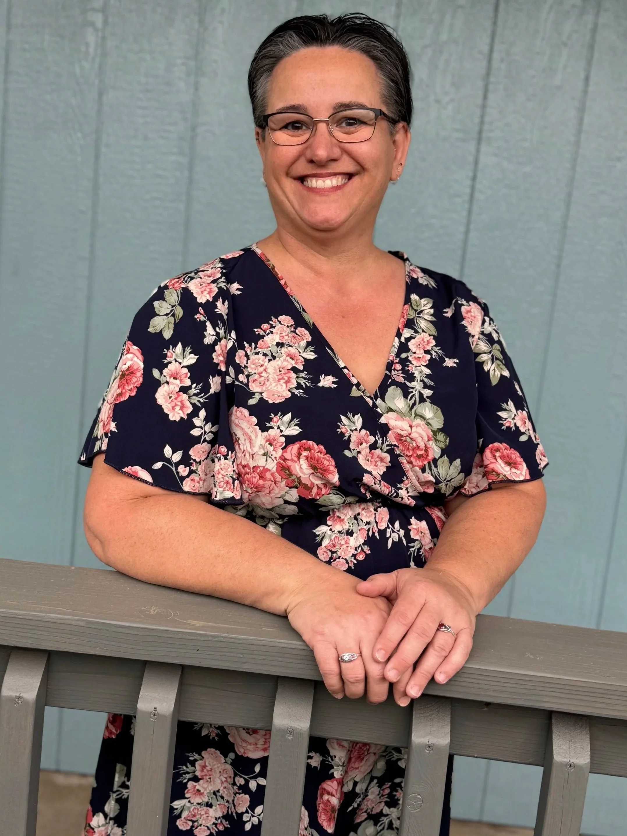 Woman with short gray hair, glasses, and a floral dress smiling while leaning on a gray wooden railing in front of a blue wall.