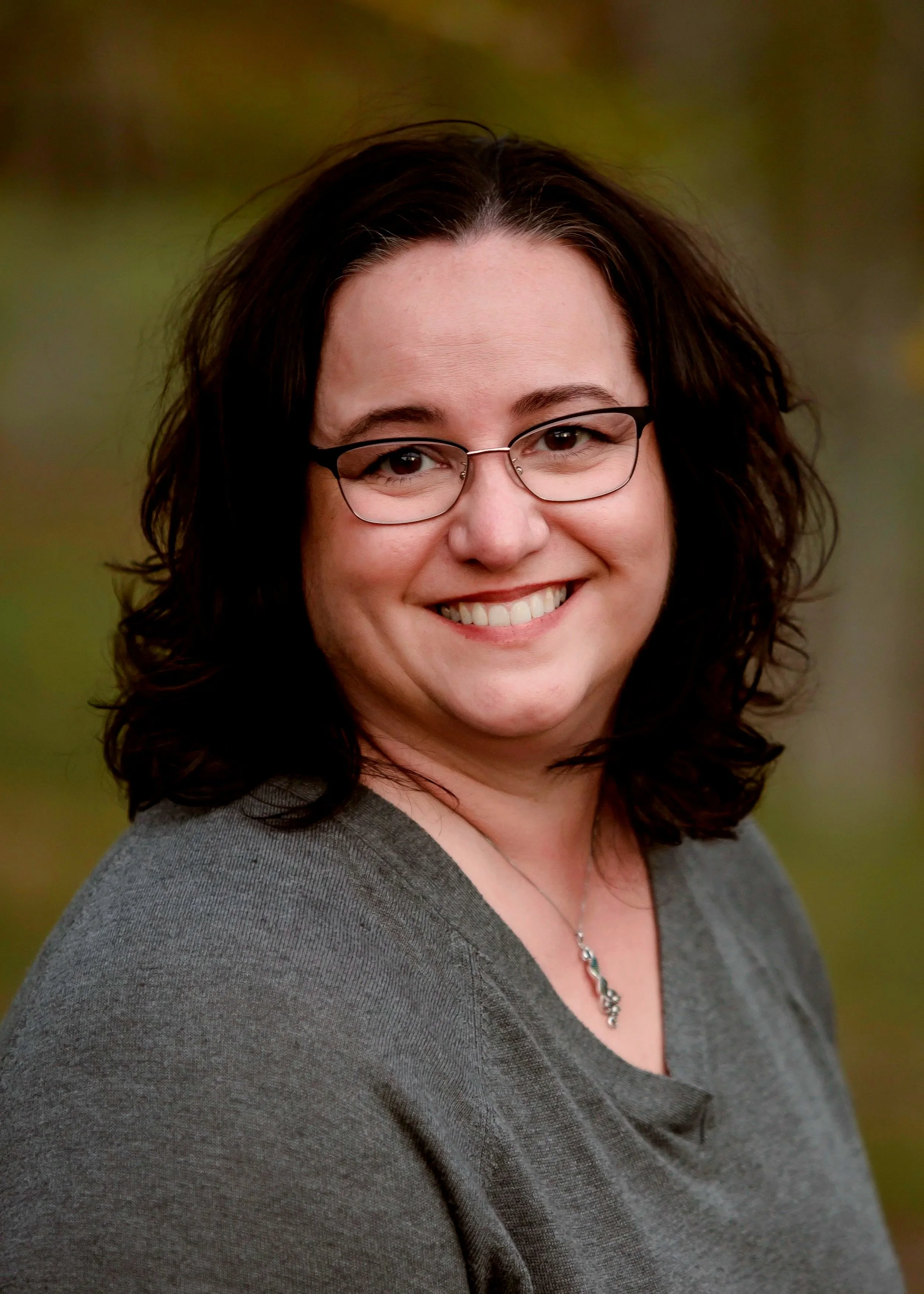 A woman with short curly brown hair, glasses, and a grey top smiling outdoors.