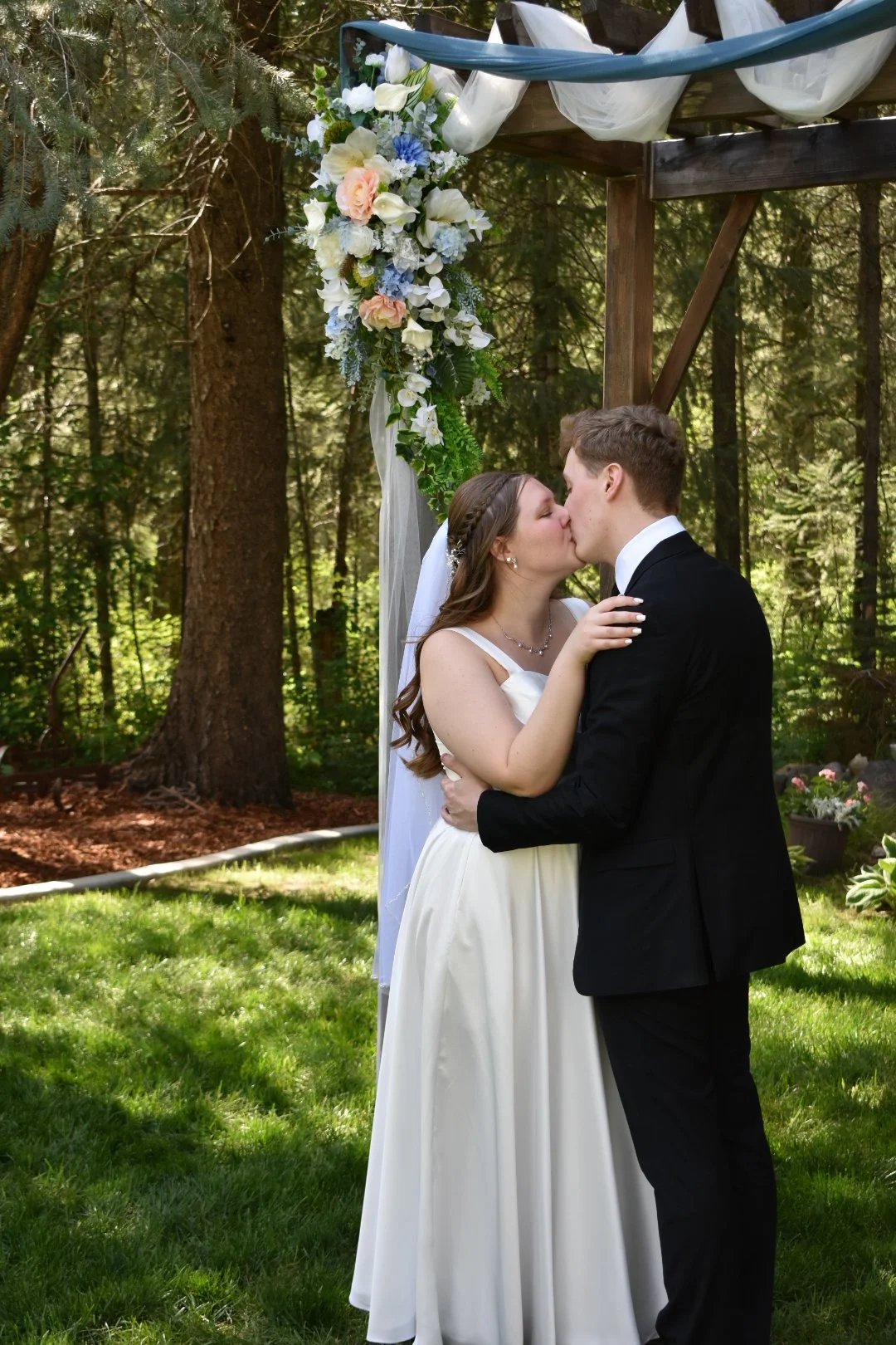 A bride and groom share a kiss and embrace during their outdoor wedding ceremony in a forest, with a floral arch above them.