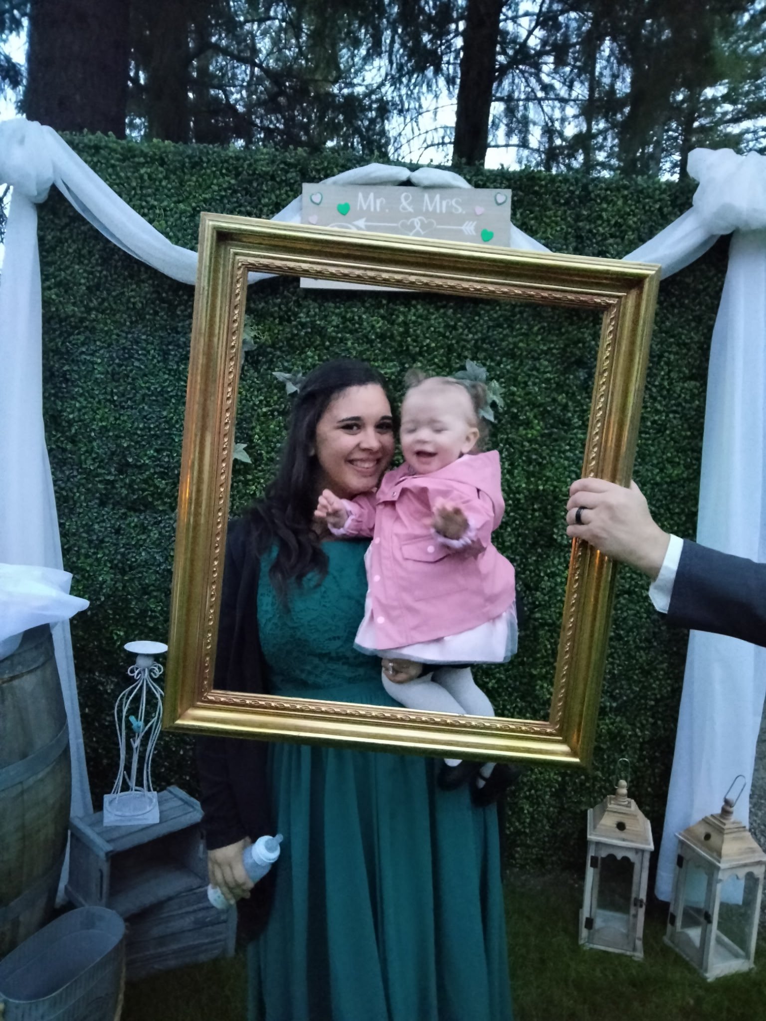 A woman holding a smiling young girl in front of a green hedge backdrop decorated with white drapes, lanterns, and a sign that reads 'Mr. & Mrs.' seen through a large picture frame.