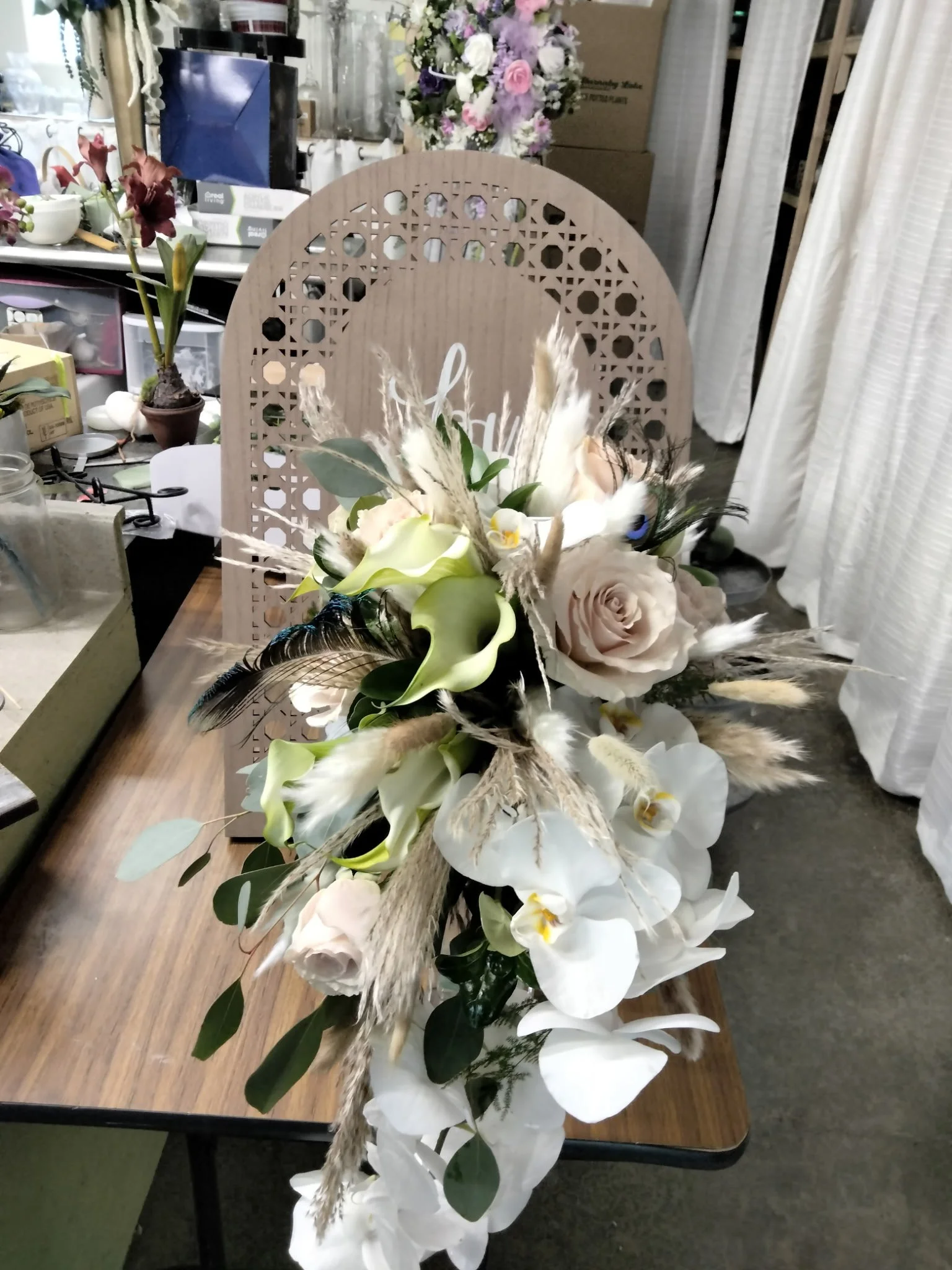 A flower arrangement with white orchids, roses, calla lilies, peacock feathers, and dried grasses on a wooden table.