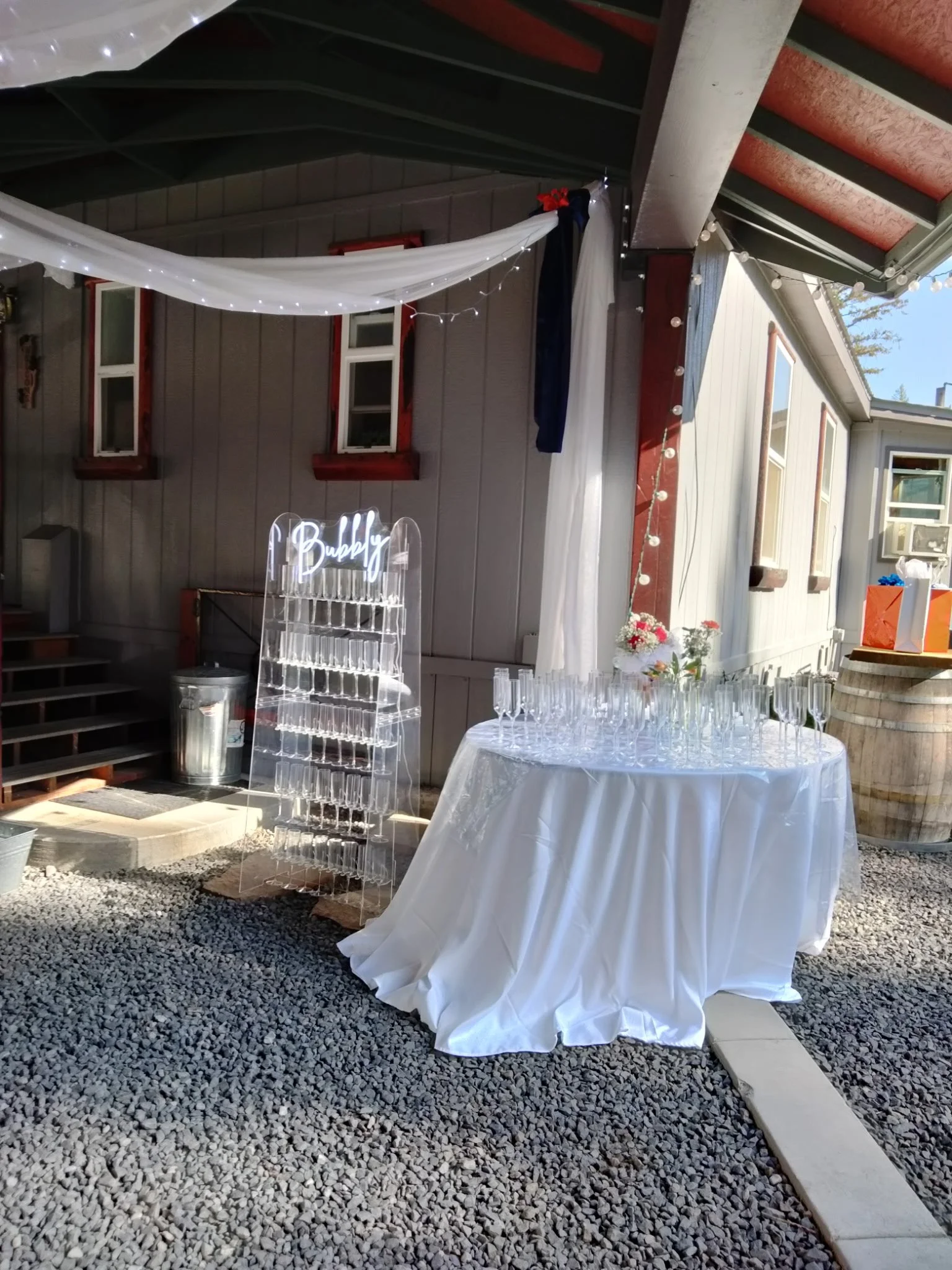 A table covered with a white tablecloth, set with many champagne glasses, a small flower arrangement, and a backlit sign that says 'Bubby,' positioned outside under a covered patio decorated with white and navy drapes and string lights.