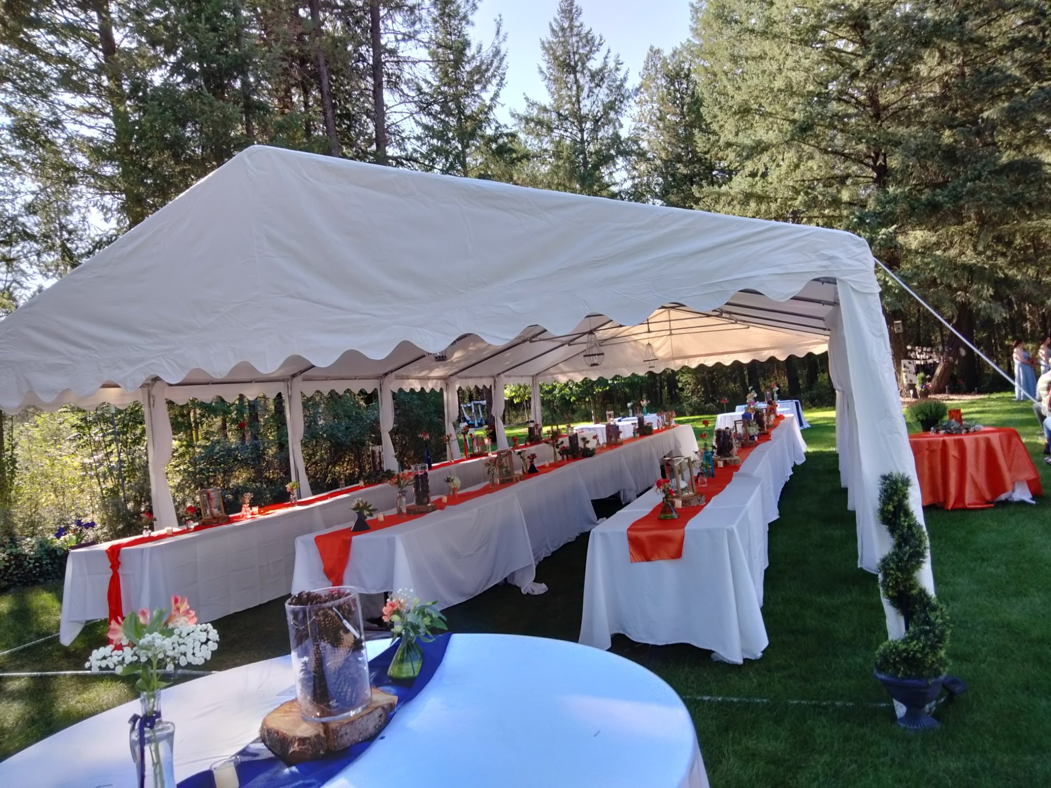 Outdoor event setup with long tables under a white canopy tent, decorated with orange table runners, small floral arrangements, and framed photographs, on a lawn surrounded by trees.