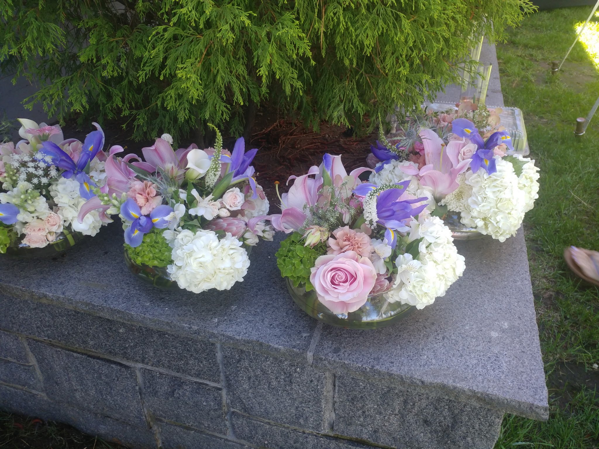 Three glass bowls filled with pink, purple, and white floral arrangements on a stone surface outdoors.