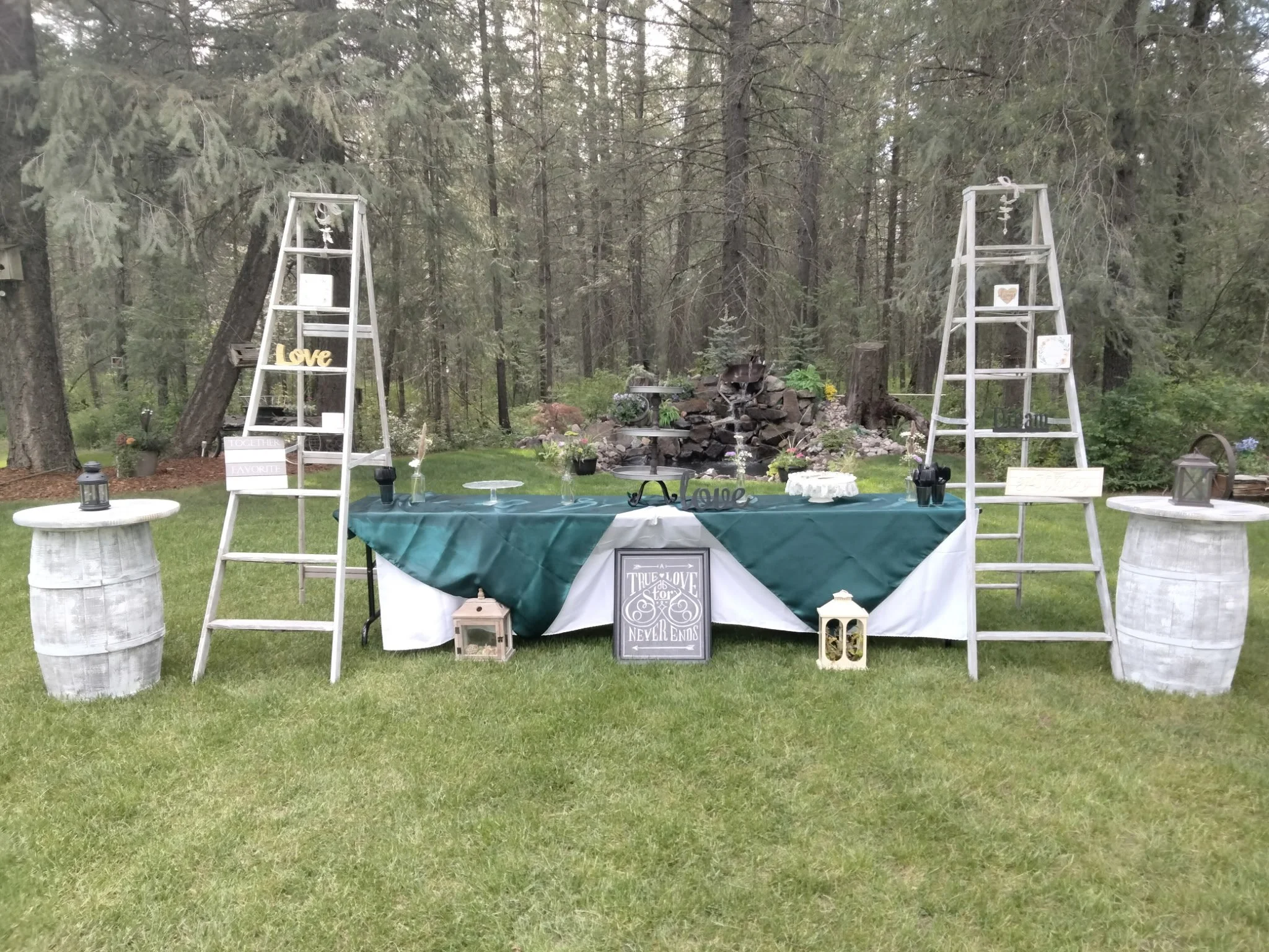 Outdoor party setup with a rectangular table covered with a green and white tablecloth, decorative lanterns, picture frames, and a centerpiece. Two white wooden ladders decorated with signs and small items are placed on either side of the table. Ther