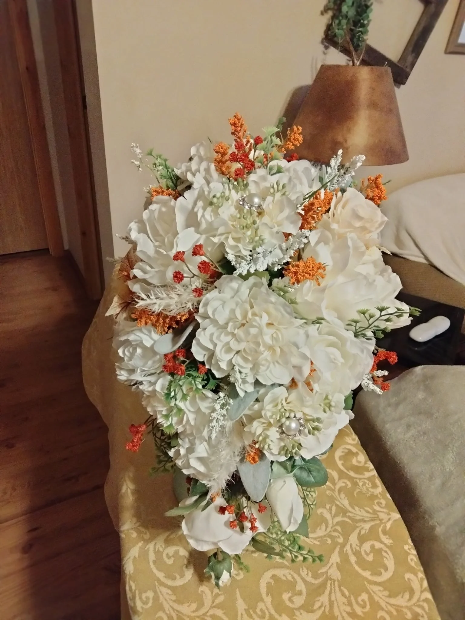 A large floral arrangement with white flowers, orange sprigs, green foliage, and faux pearl accents, placed on a patterned tablecloth near a lamp and a beige armchair.